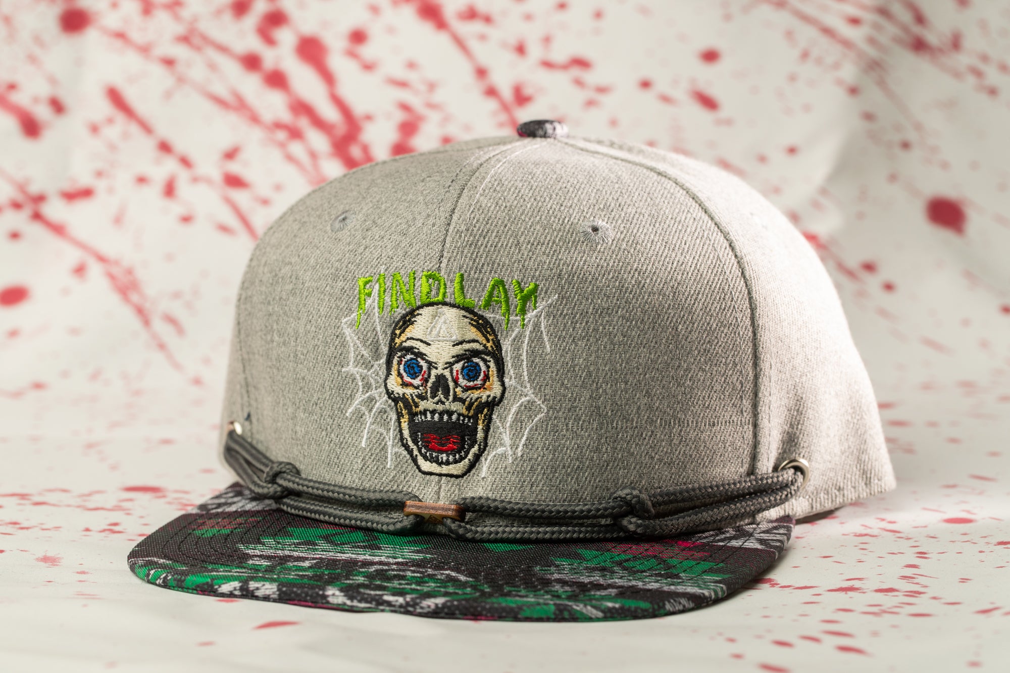 A gray baseball cap with a skull design and the text "FIND LAY" printed on it, set against a background with red splatters.
