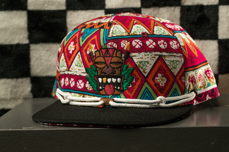 A colorful, patterned baseball cap with a tiki mask design on the front, displayed on a black surface against a checkered background.