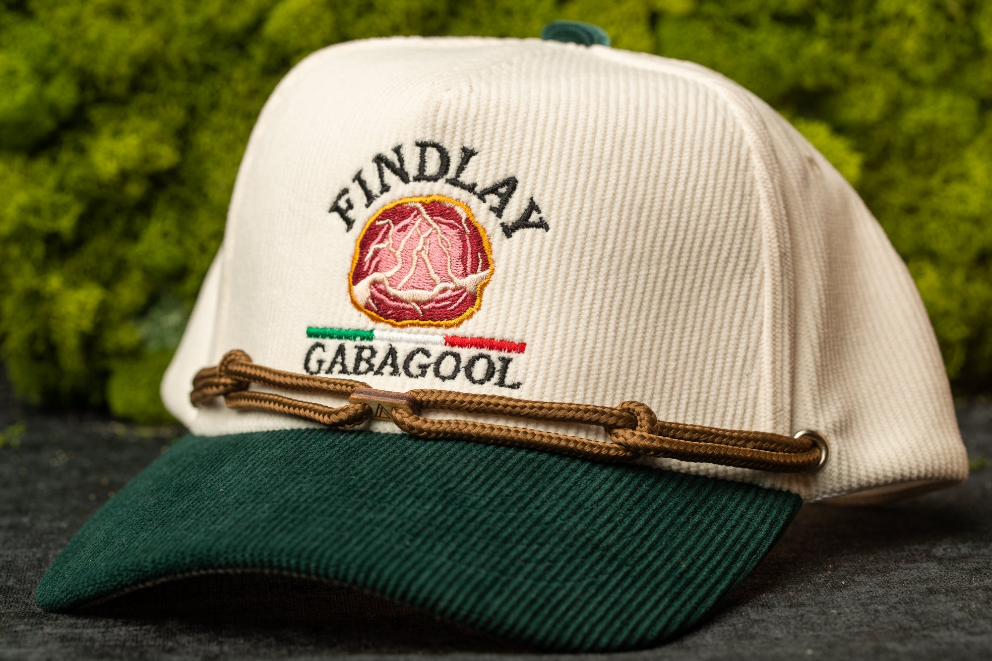 A white baseball cap with a green brim and a brown leather strap, featuring a logo on the front.