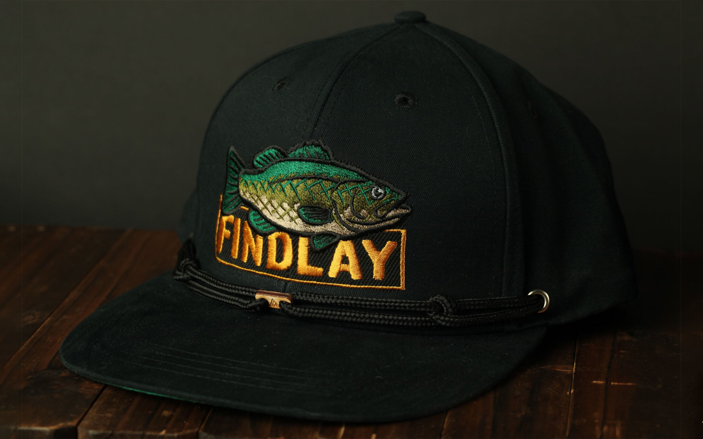 A black baseball cap with a fish embroidered on it, sitting on a wooden surface.