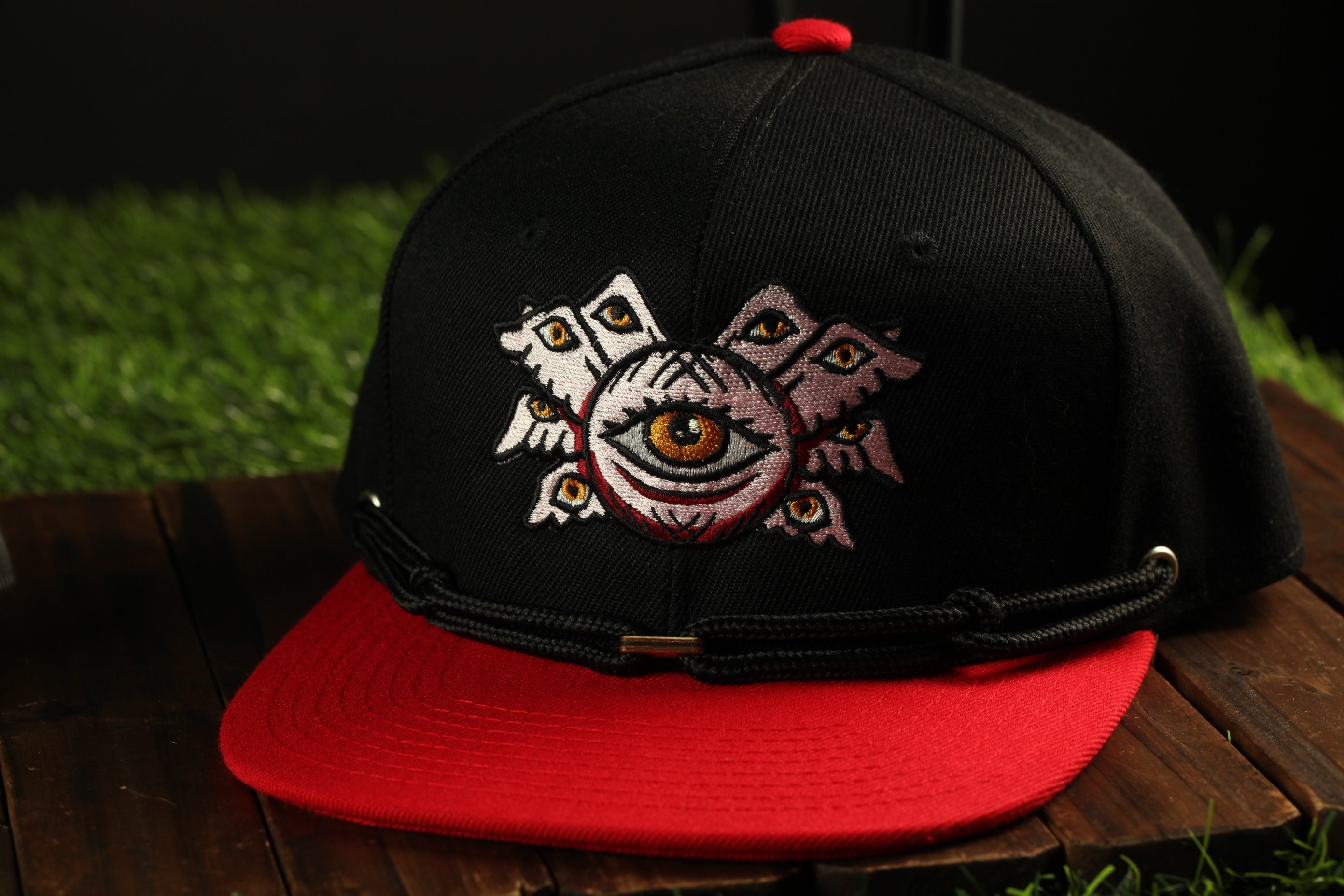 A black baseball cap with a red brim, featuring an embroidered design of a large eye surrounded by smaller eyes, is displayed on a wooden surface.