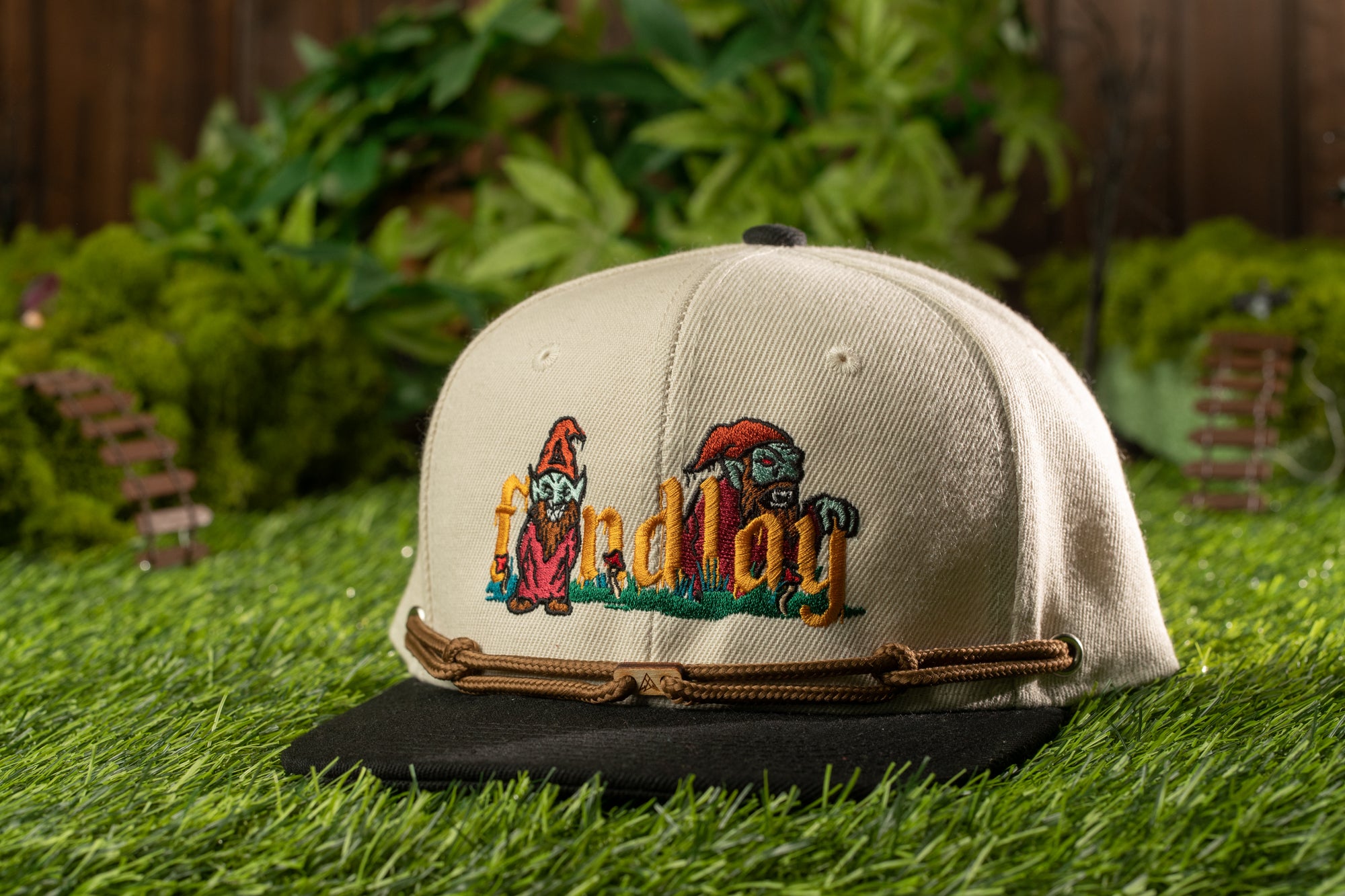A beige baseball cap with a cartoonish design and the word "lay" printed on it, placed on a grassy surface with a small wooden structure in the background.