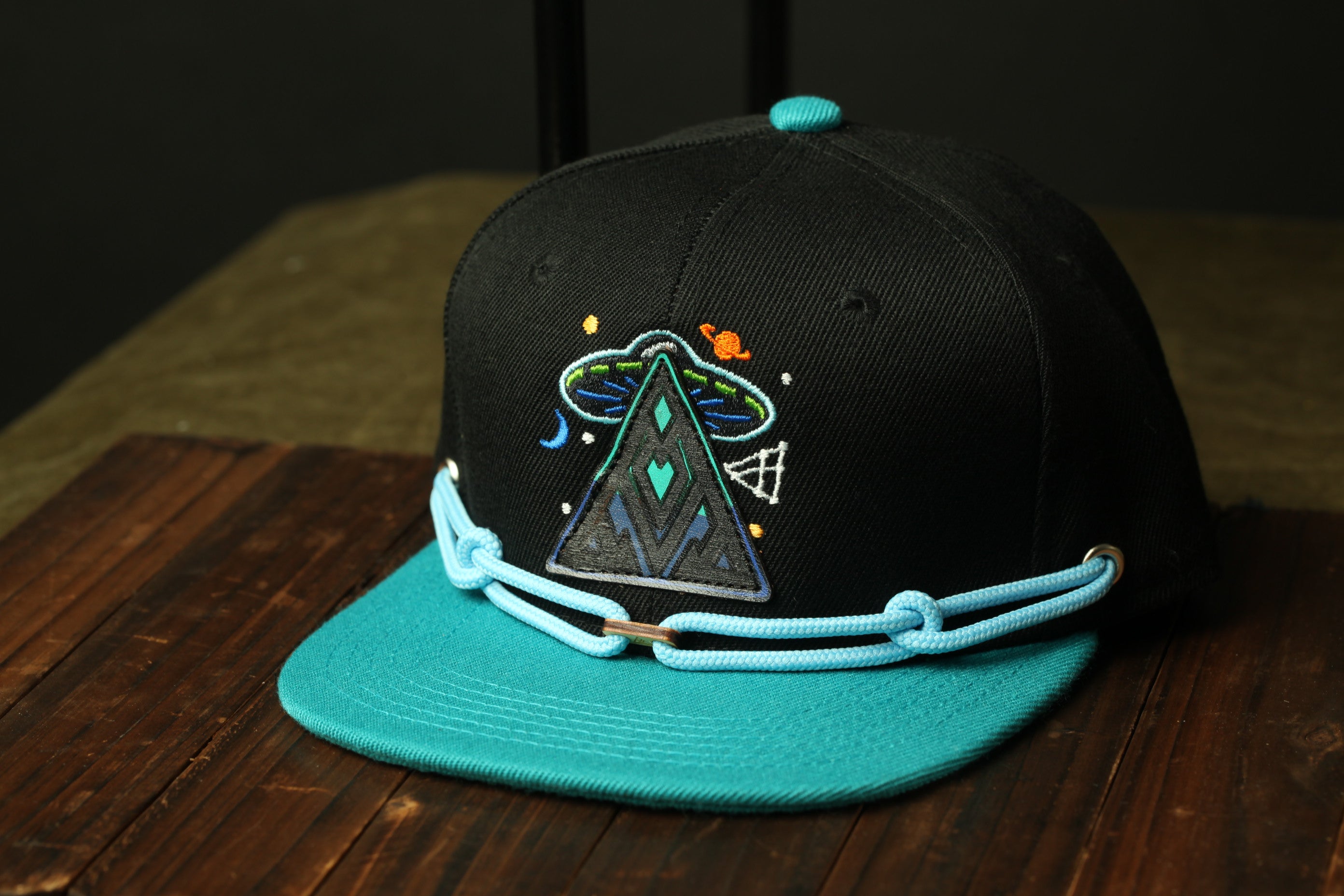A black baseball cap with a teal brim and a colorful alien design on the front, sitting on a wooden surface.