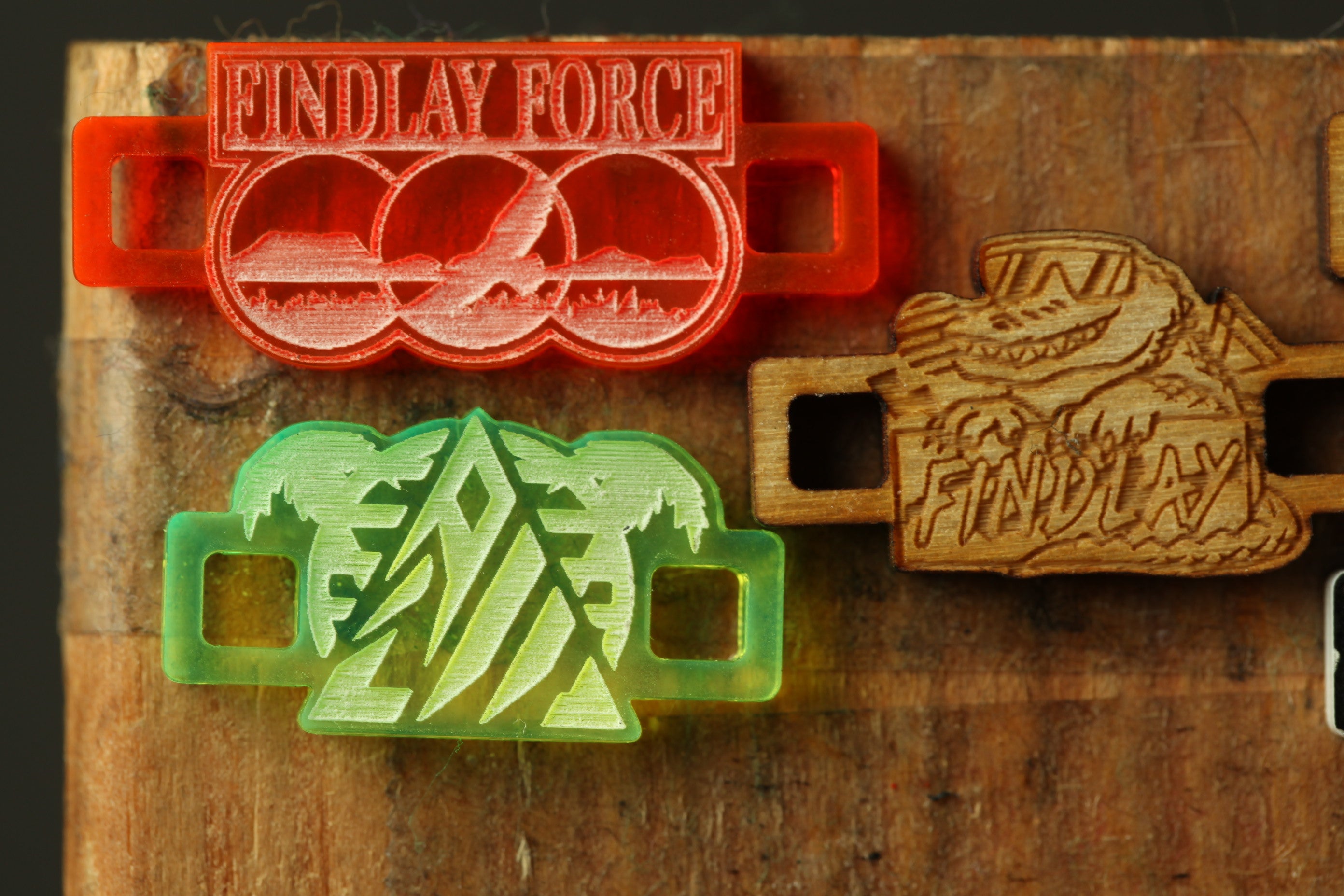 three different objects, including a red and orange object with the text "FINDLAY FORCE" on it, a green object with a triangle design, and a wooden object with the text "FINDLAY" carved into it.
