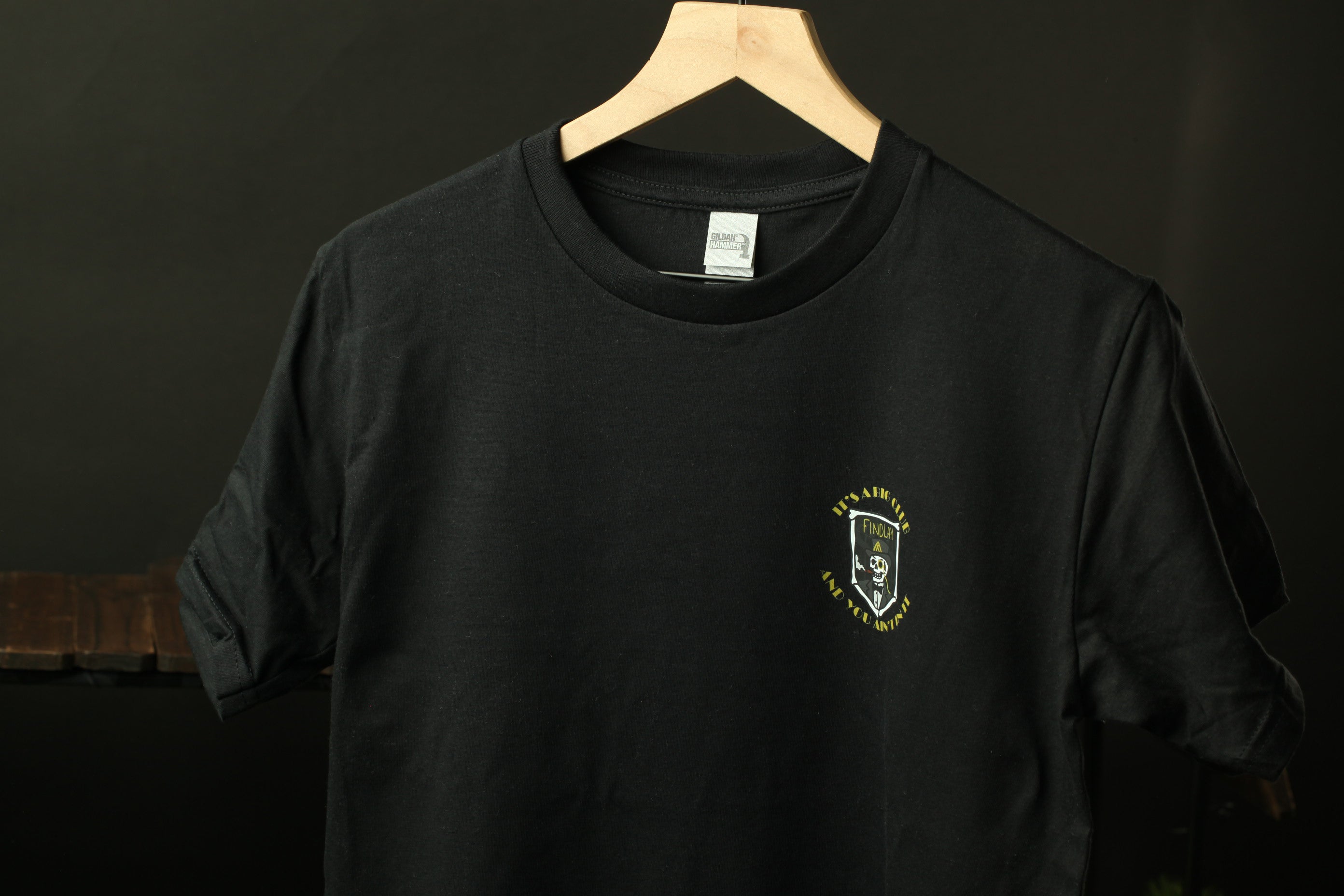 A black t-shirt with a small logo on the front, hanging on a wooden hanger against a dark background.