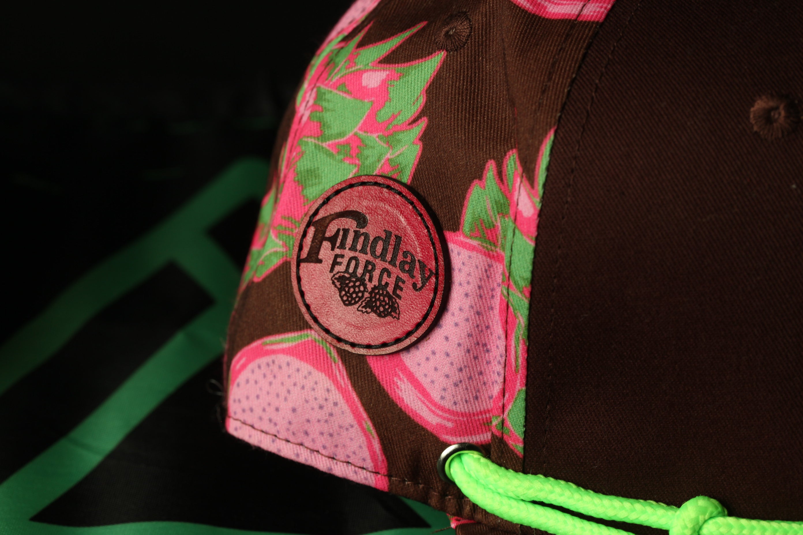 A close-up view of a brown baseball cap with a pink and green floral pattern, featuring a circular patch with the text "Fundlay Force" on it.