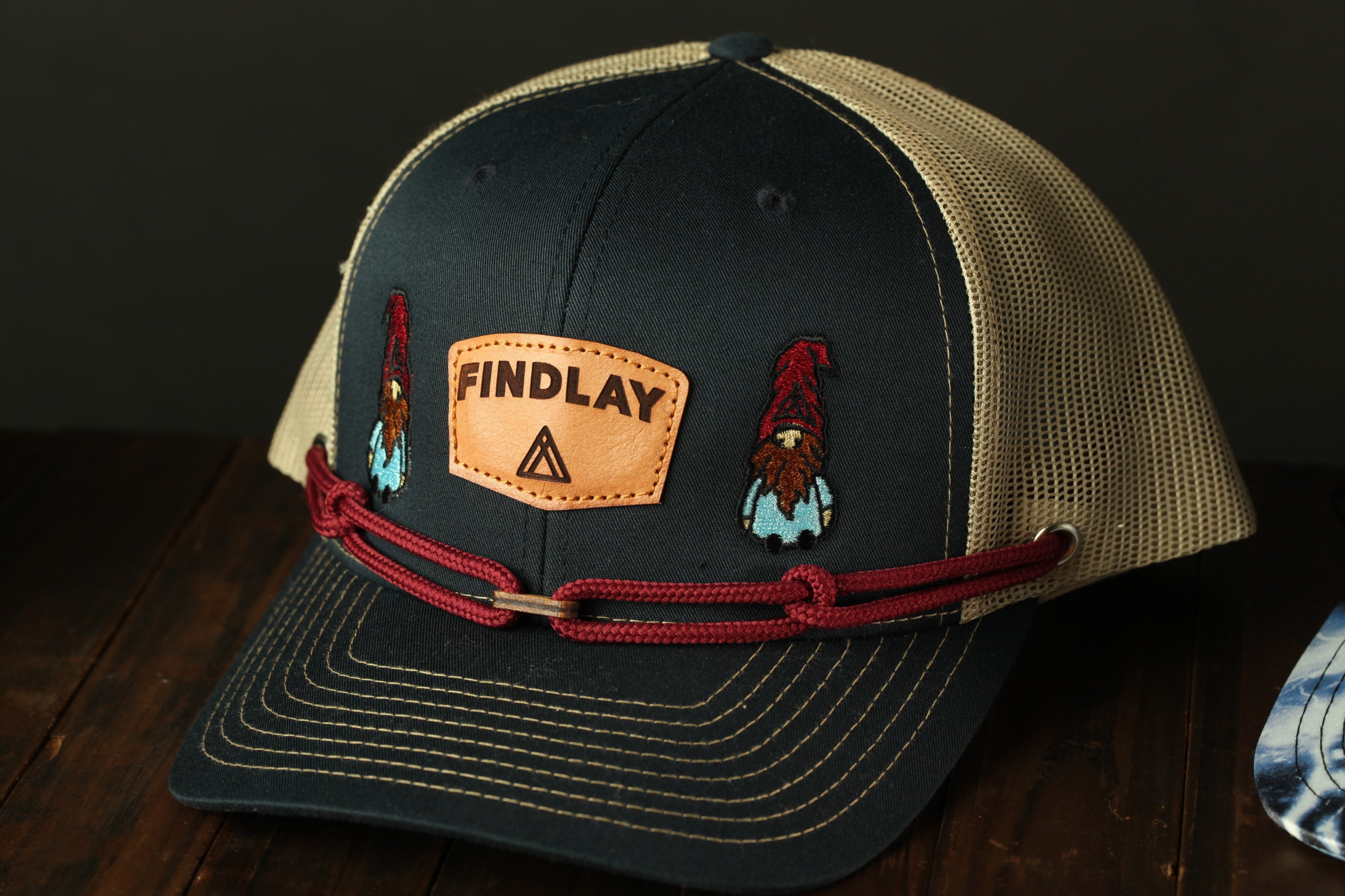 A black baseball cap with a mesh panel and a red braided strap, featuring a patch with the word "FINDLAY" and two gnome-like figures on it.