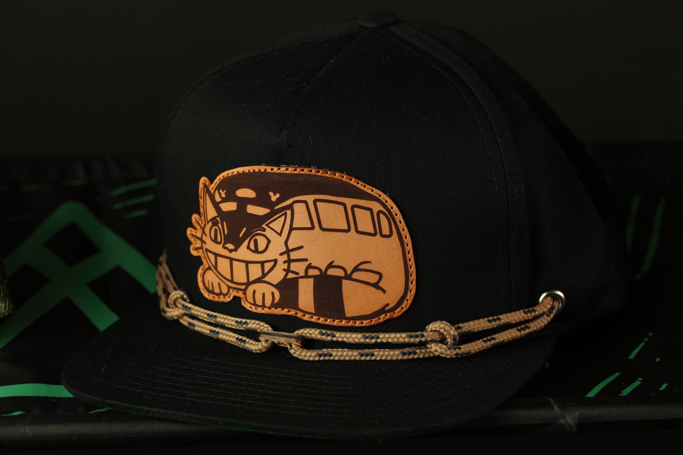A black baseball cap with a cat design on the front, featuring a smiling cat and a rope chain.
