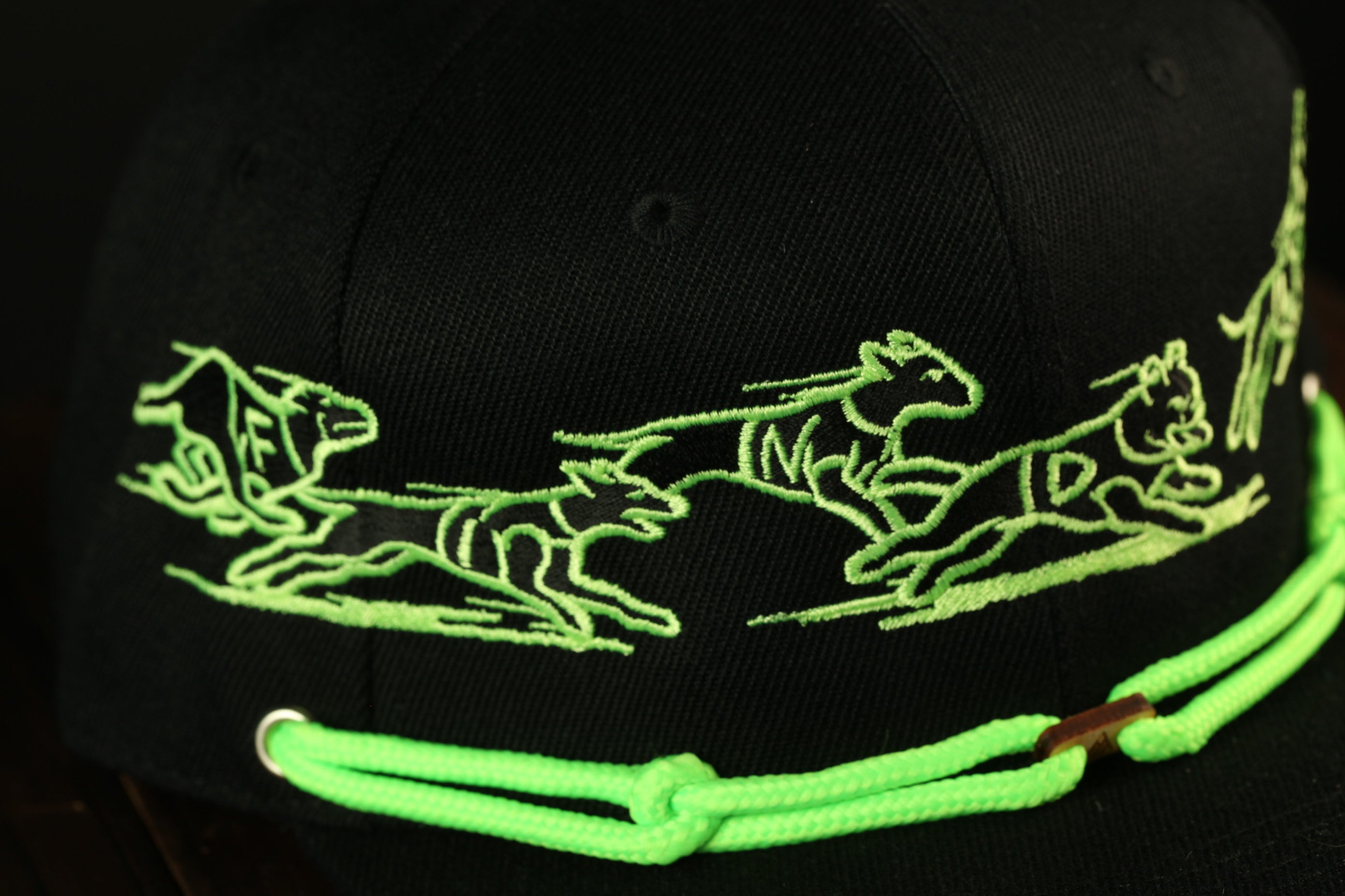 A close-up view of a black baseball cap with a green rope strap and a design of three stylized animal figures on the front.