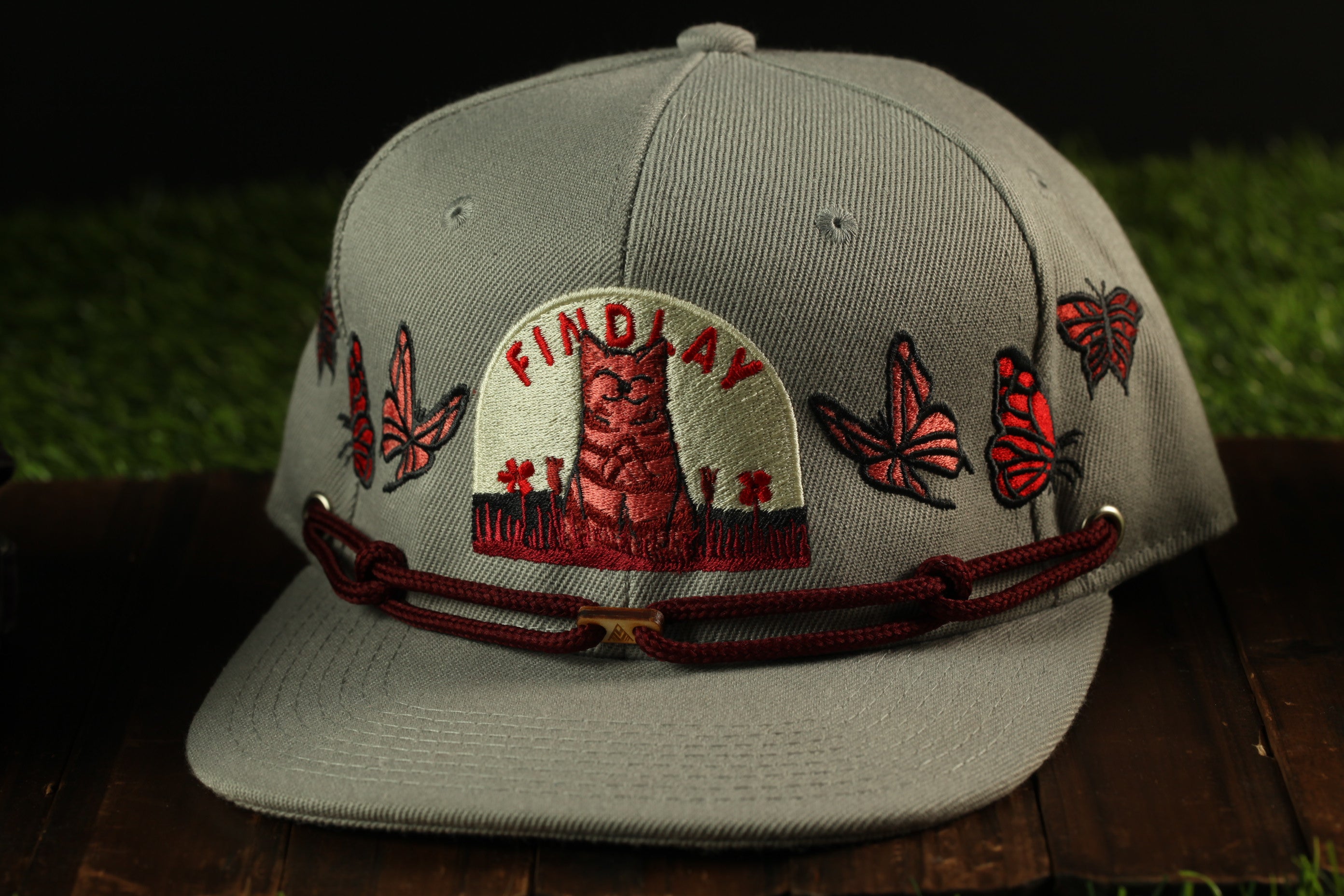 A gray baseball cap with a red and white embroidered design, featuring a red and white cat and butterflies, and the word "FINDlay" on the front.
