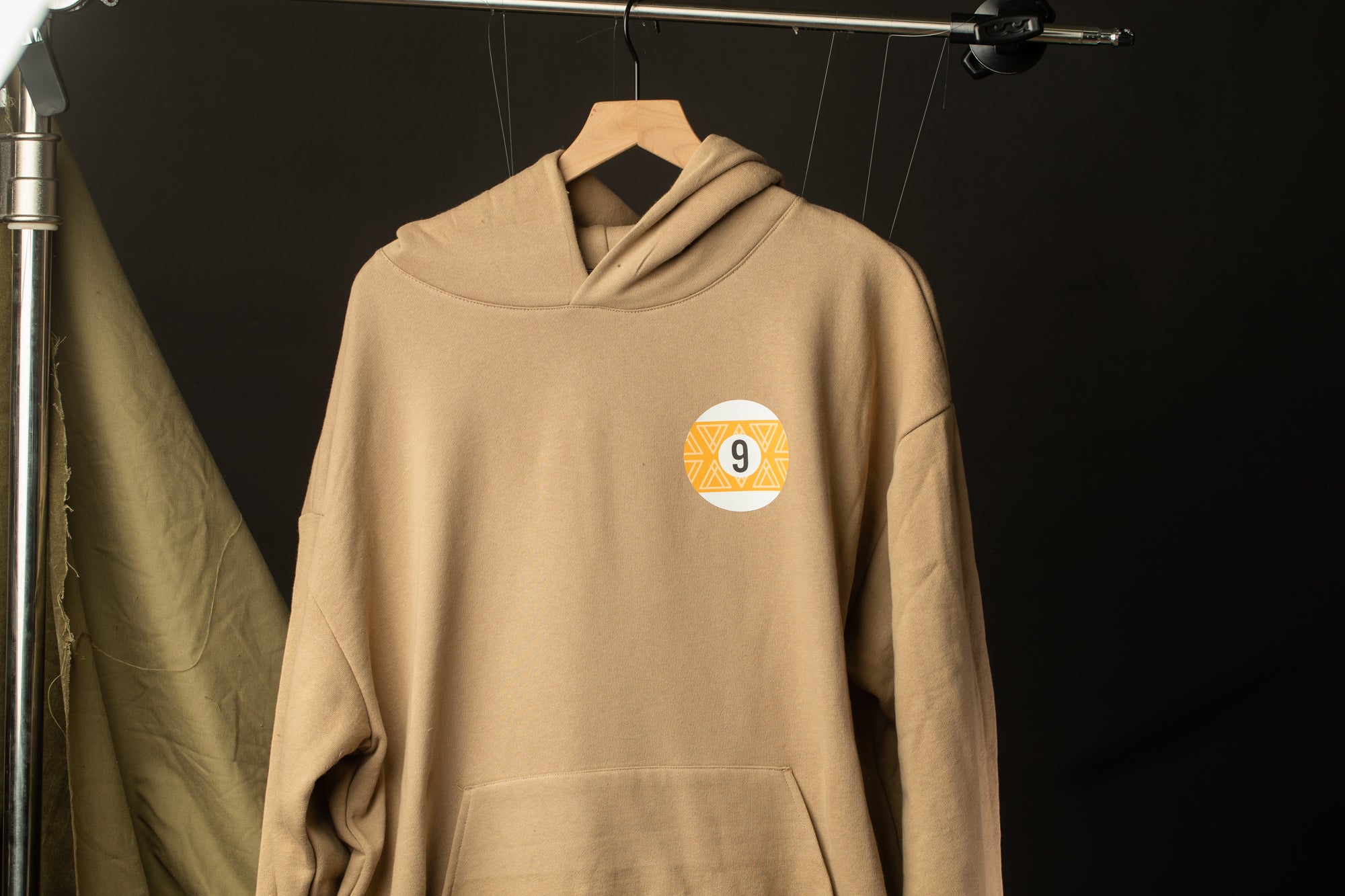 A beige hoodie with a number 9 on the front is hanging on a clothing rack against a dark background.