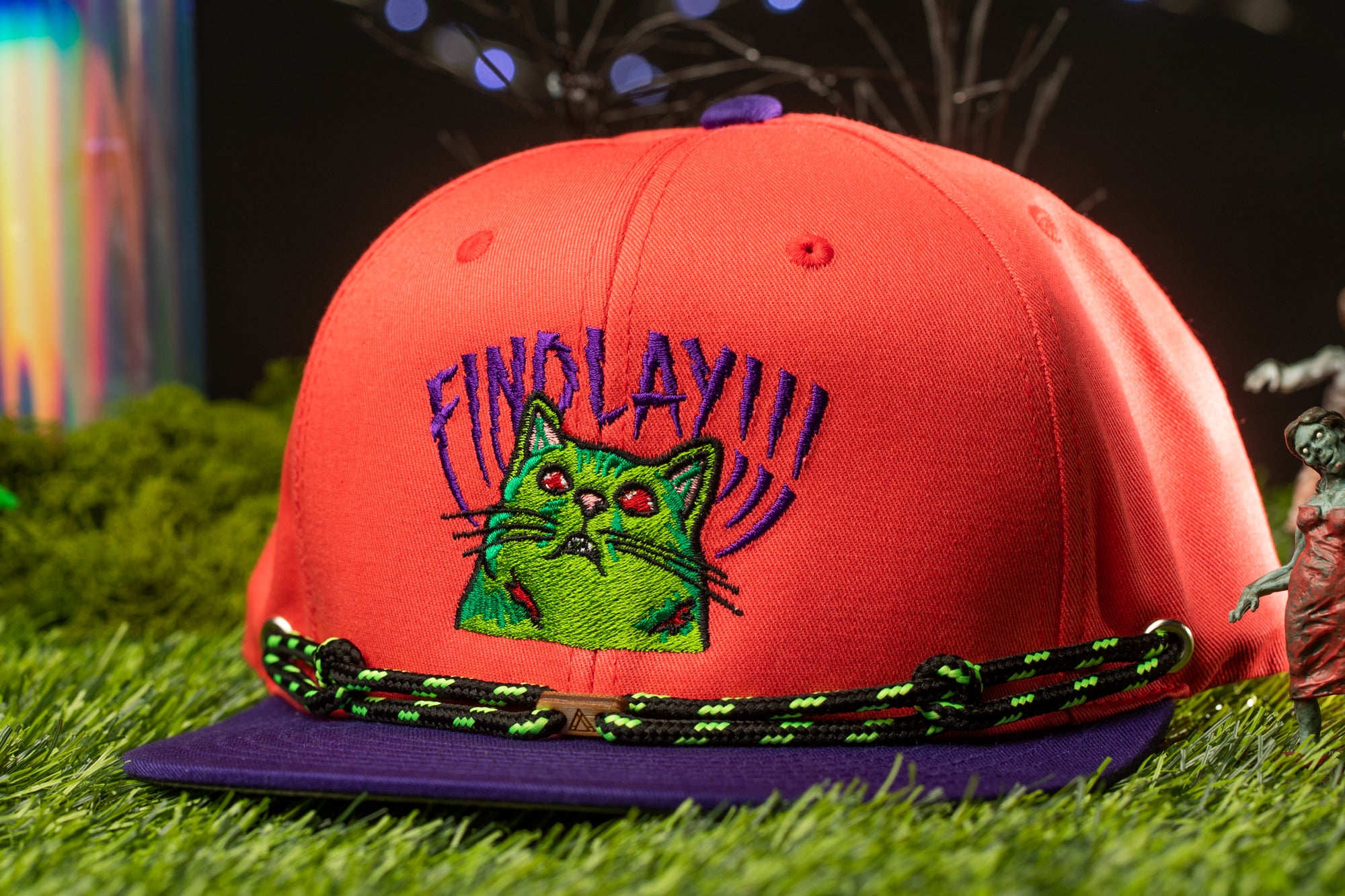 A red baseball cap with a green cat embroidered on it, sitting on a grassy surface.