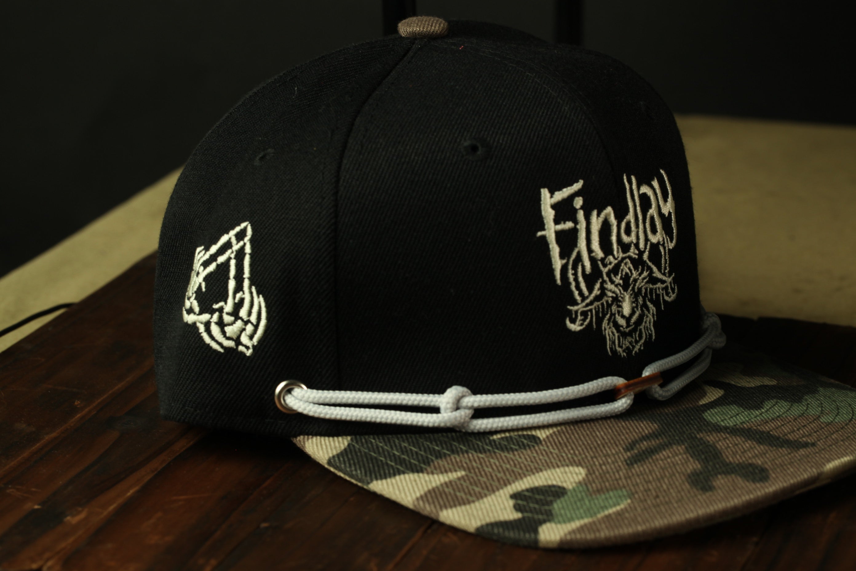 A black baseball cap with a camouflage pattern and the word "Findlay" printed on it, sitting on a wooden surface.