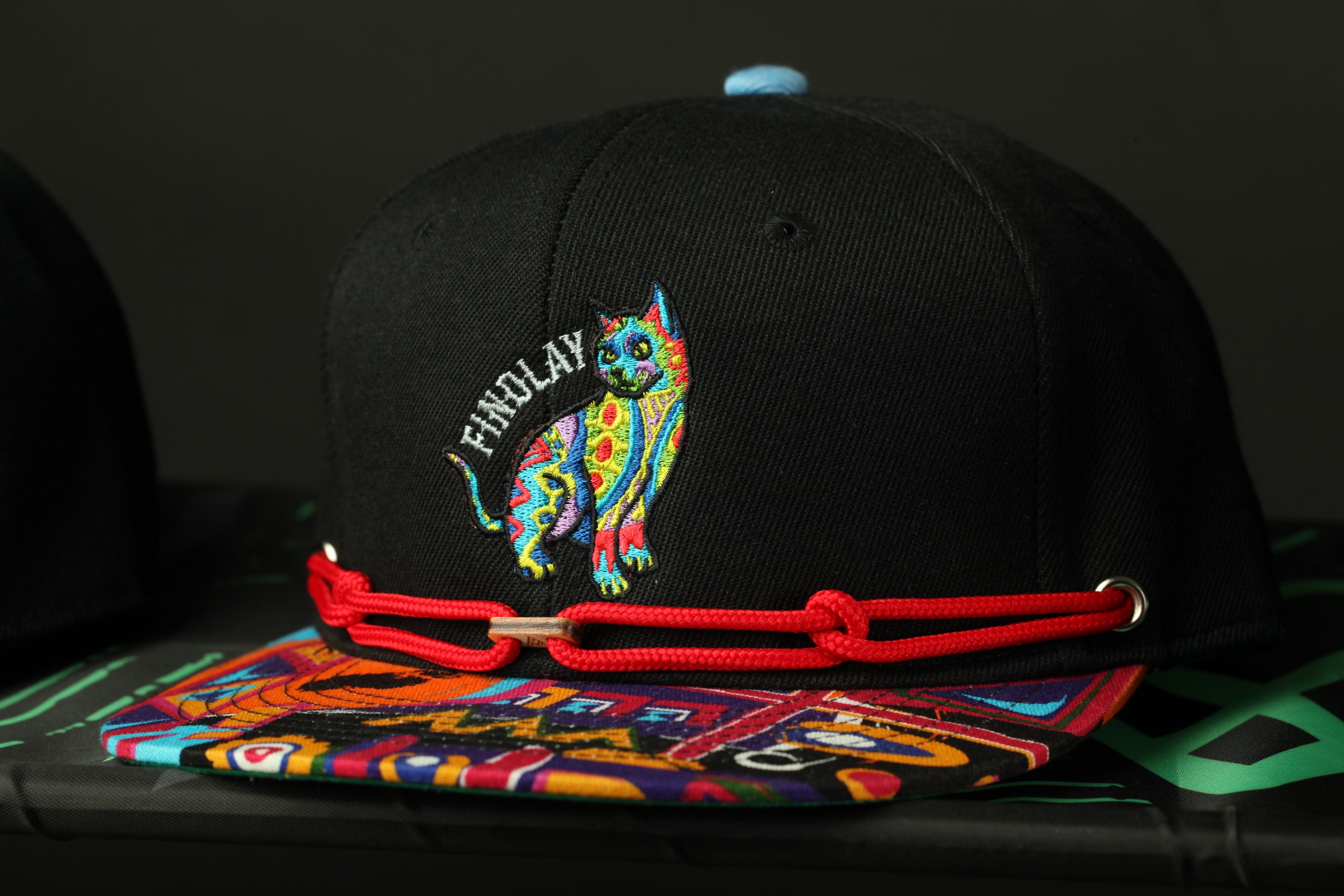 A black baseball cap with a colorful, embroidered cat design on it, and a red string or strap attached to the front.