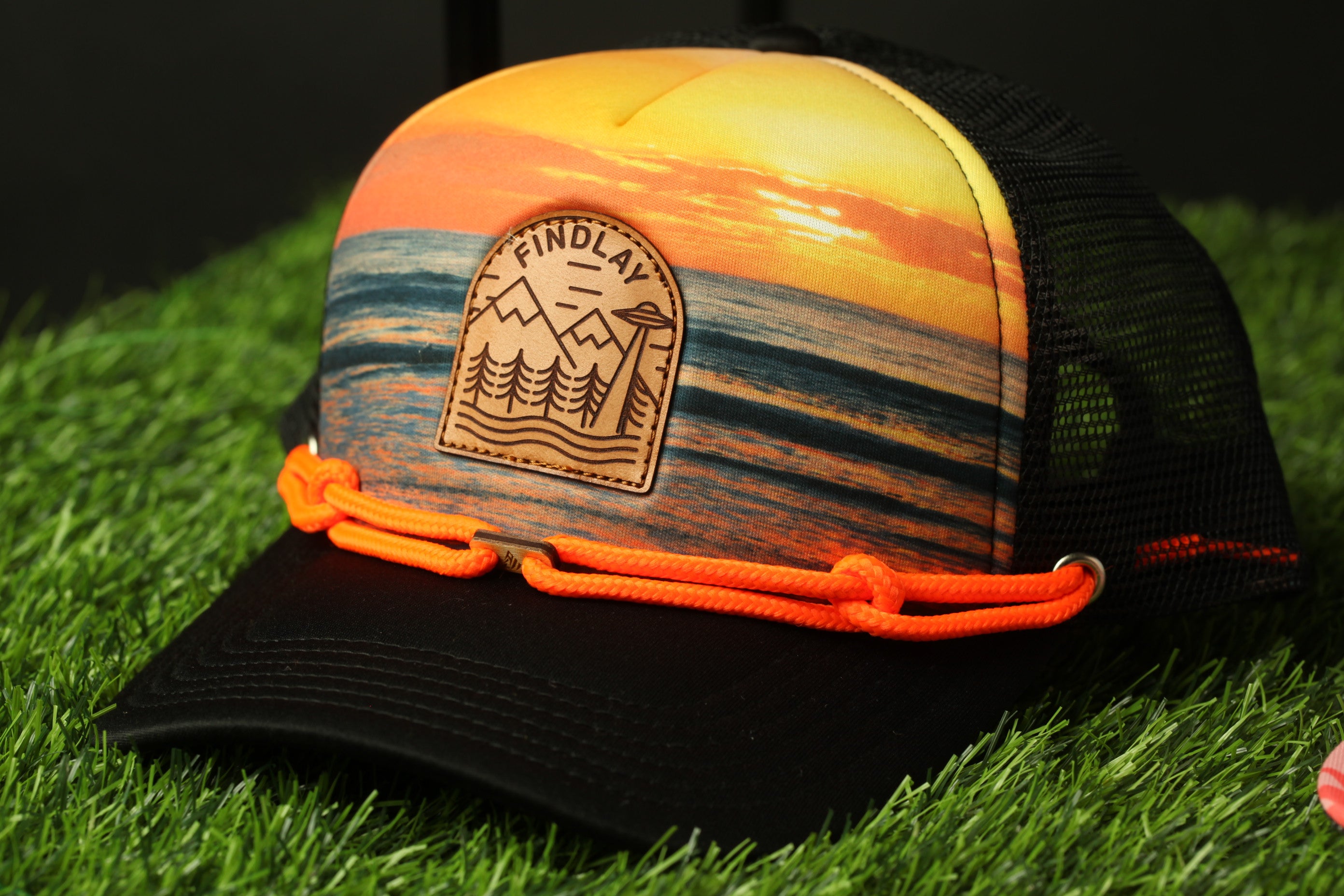 A colorful baseball cap with a printed design of a sunset landscape, featuring mountains and trees, is shown on a grassy surface.