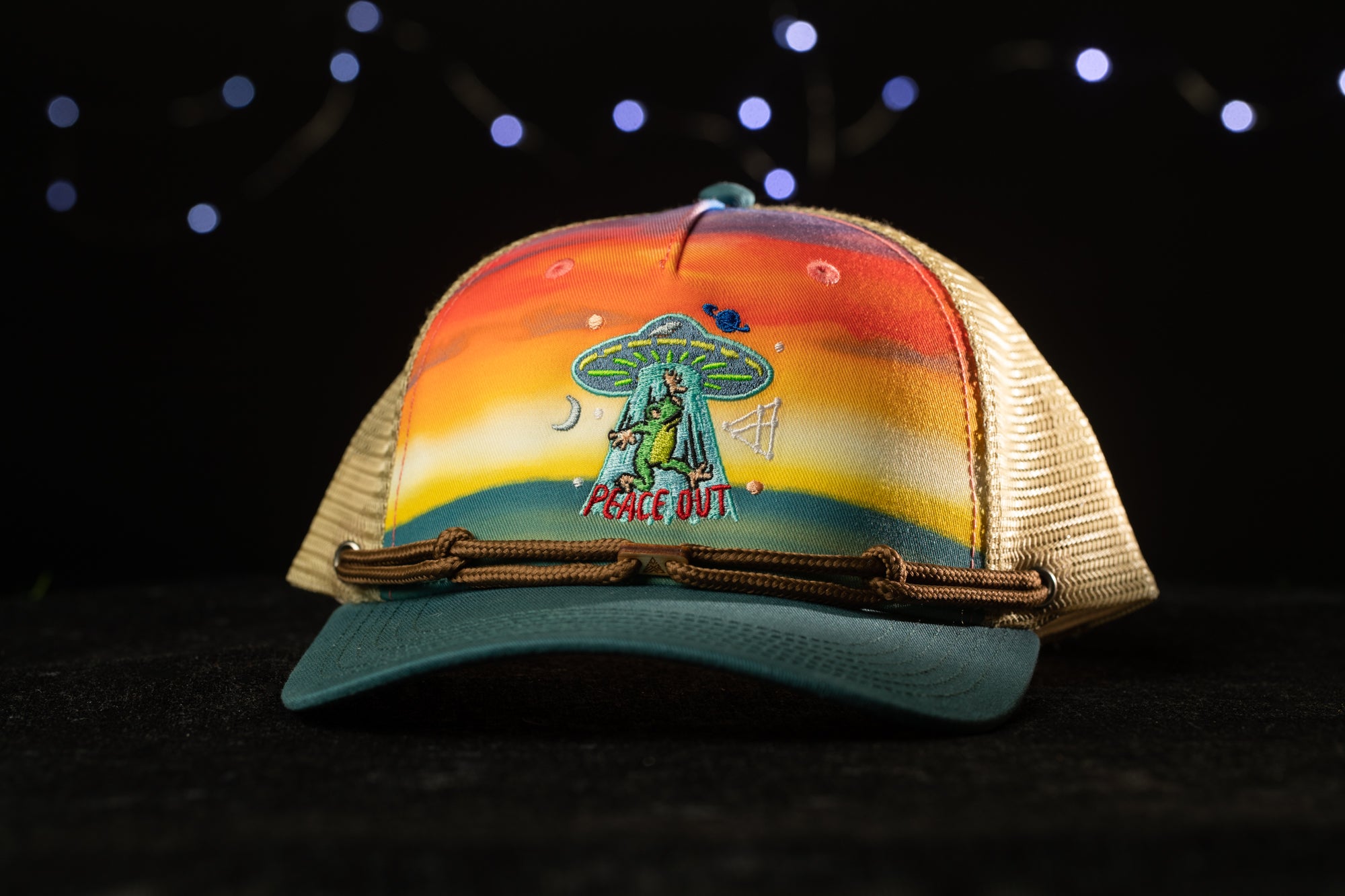 A colorful baseball cap with a graphic of an alien and the text "Peace Out" on it, set against a dark background with twinkling lights.