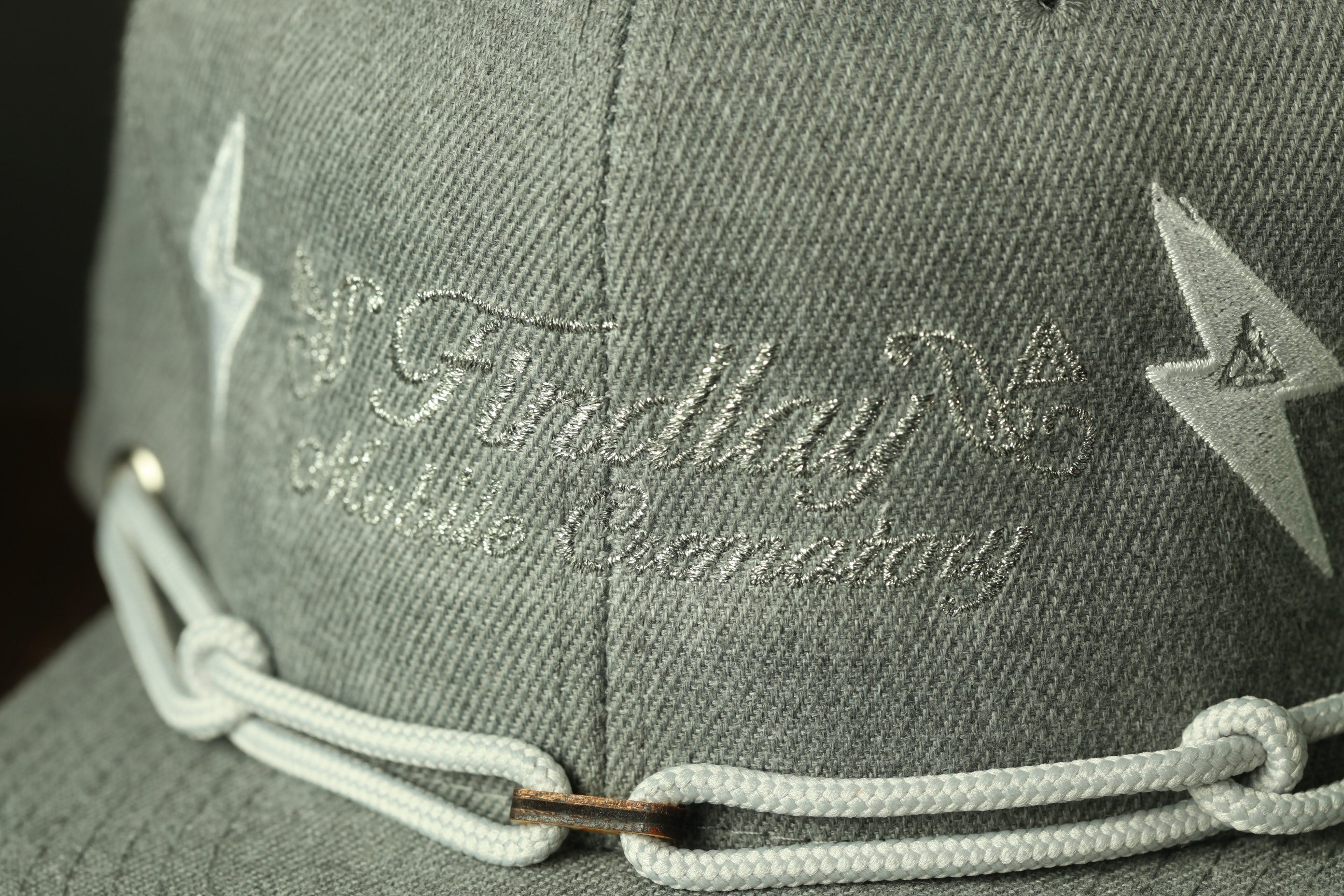 A close-up view of a gray fabric, possibly a hat or cap, with a white string or chain attached to it. The text embroidered on the fabric appears to be a name or brand, but it is not possible to provide a detailed description of the text due to the limited information provided.