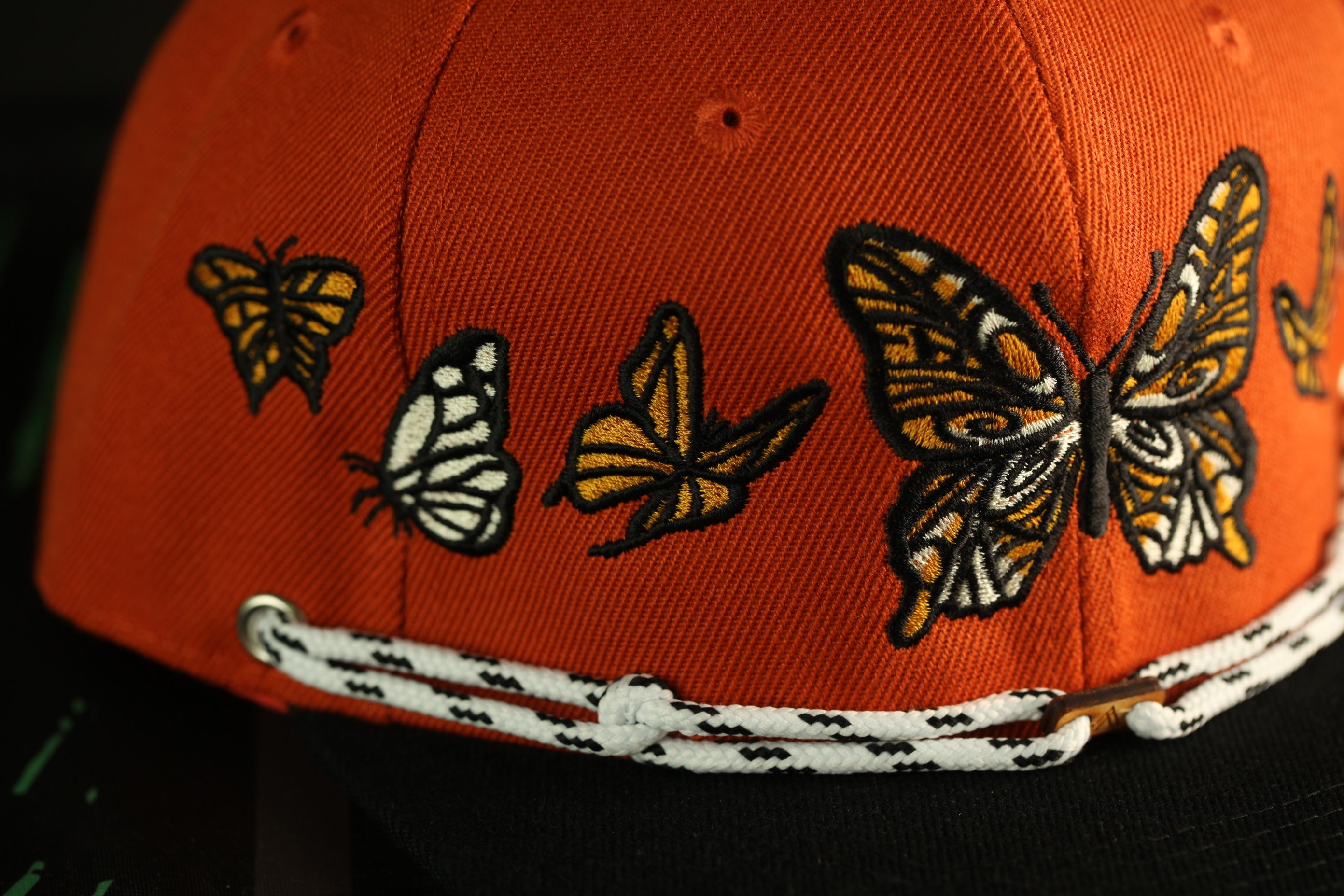 a close-up view of a red baseball cap with a white and black rope strap. The cap has a row of embroidered butterflies on the front, with one large butterfly in the center and smaller butterflies flanking it on either side.