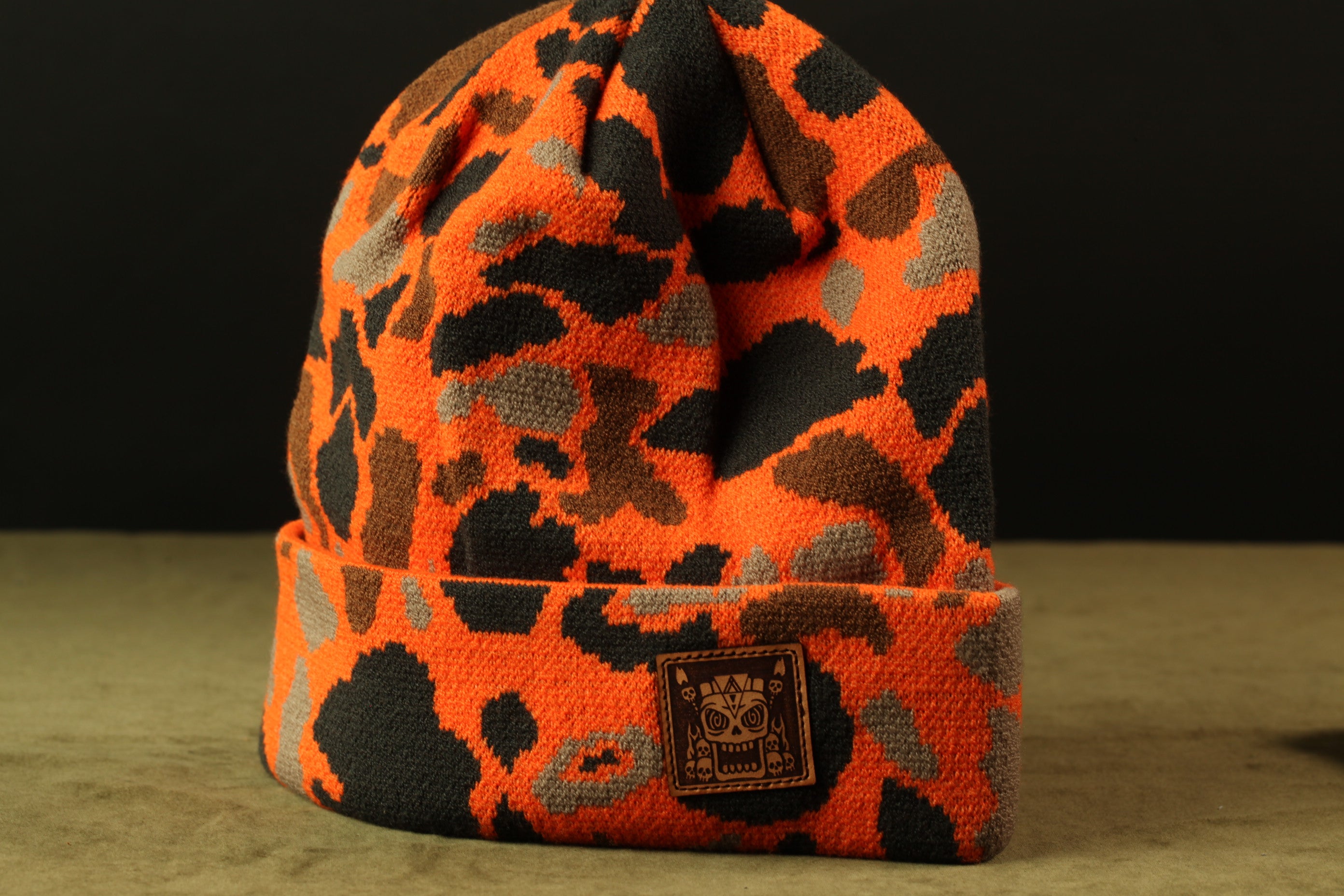 An orange and black camouflage beanie with a small patch on the front displaying a skull and crossbones symbol.