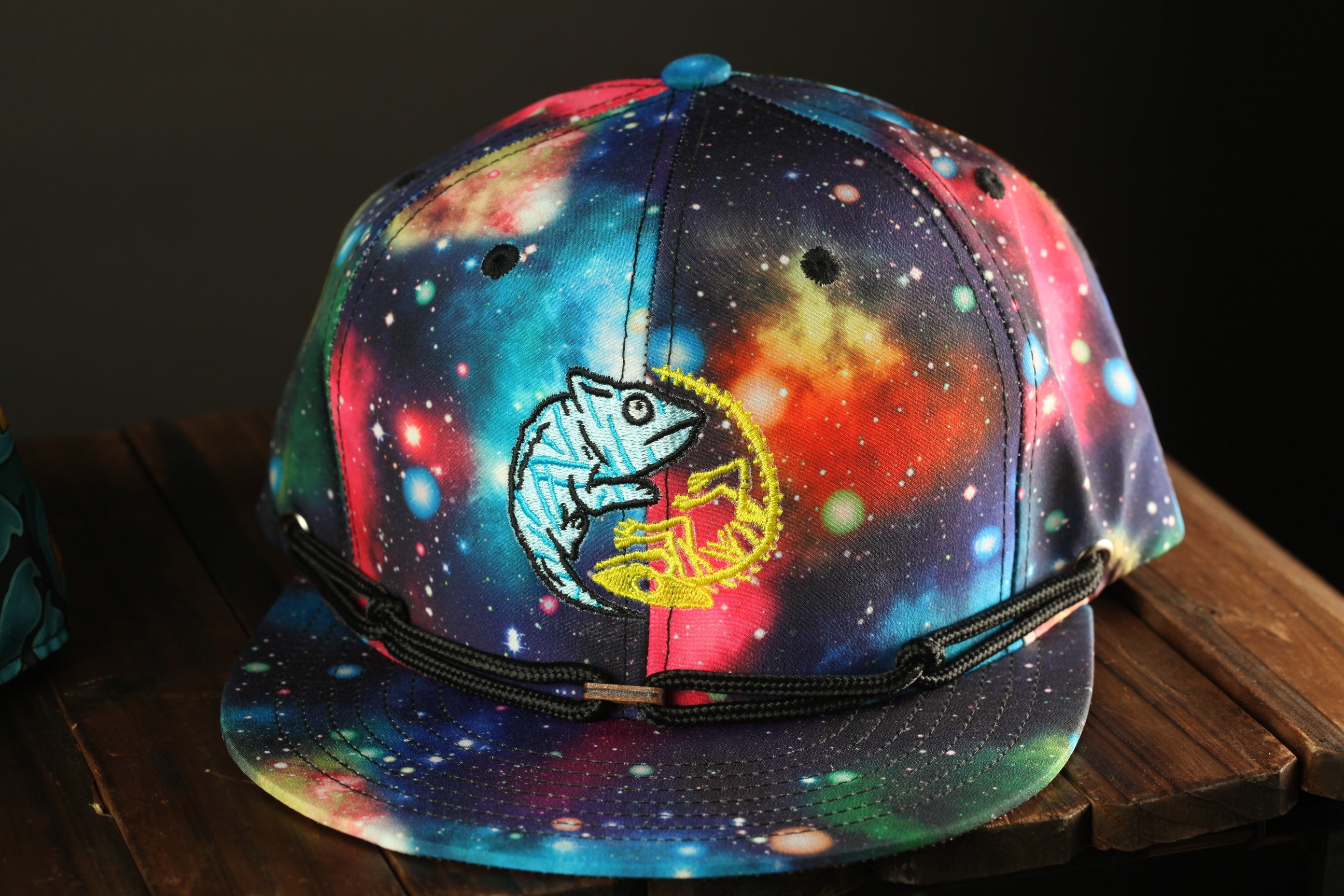A colorful, galaxy-themed baseball cap with a chameleon design, displayed on a wooden surface.