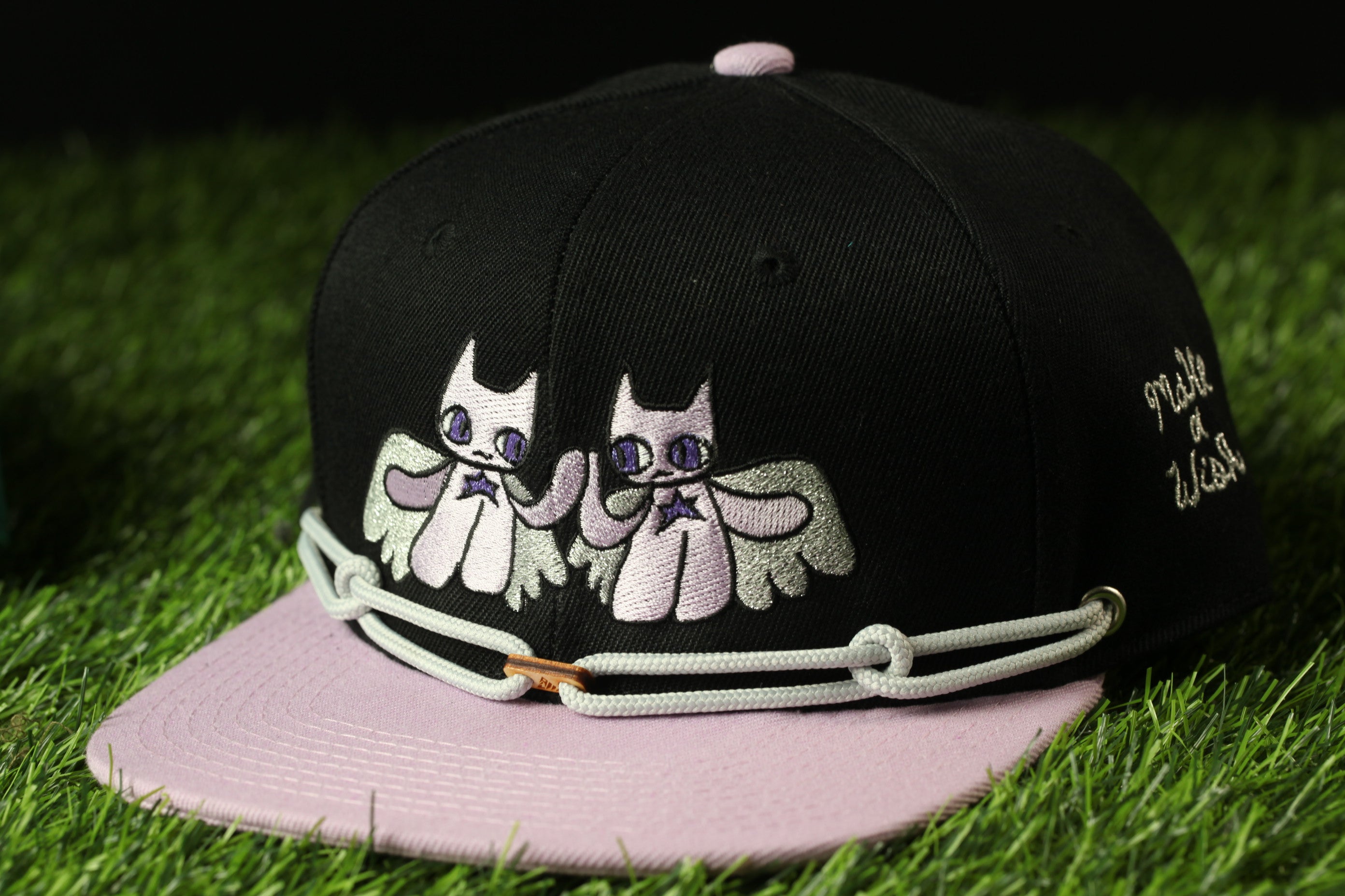 A black baseball cap with a pink brim and a design of two cartoon cats on the front, sitting on a grassy surface.