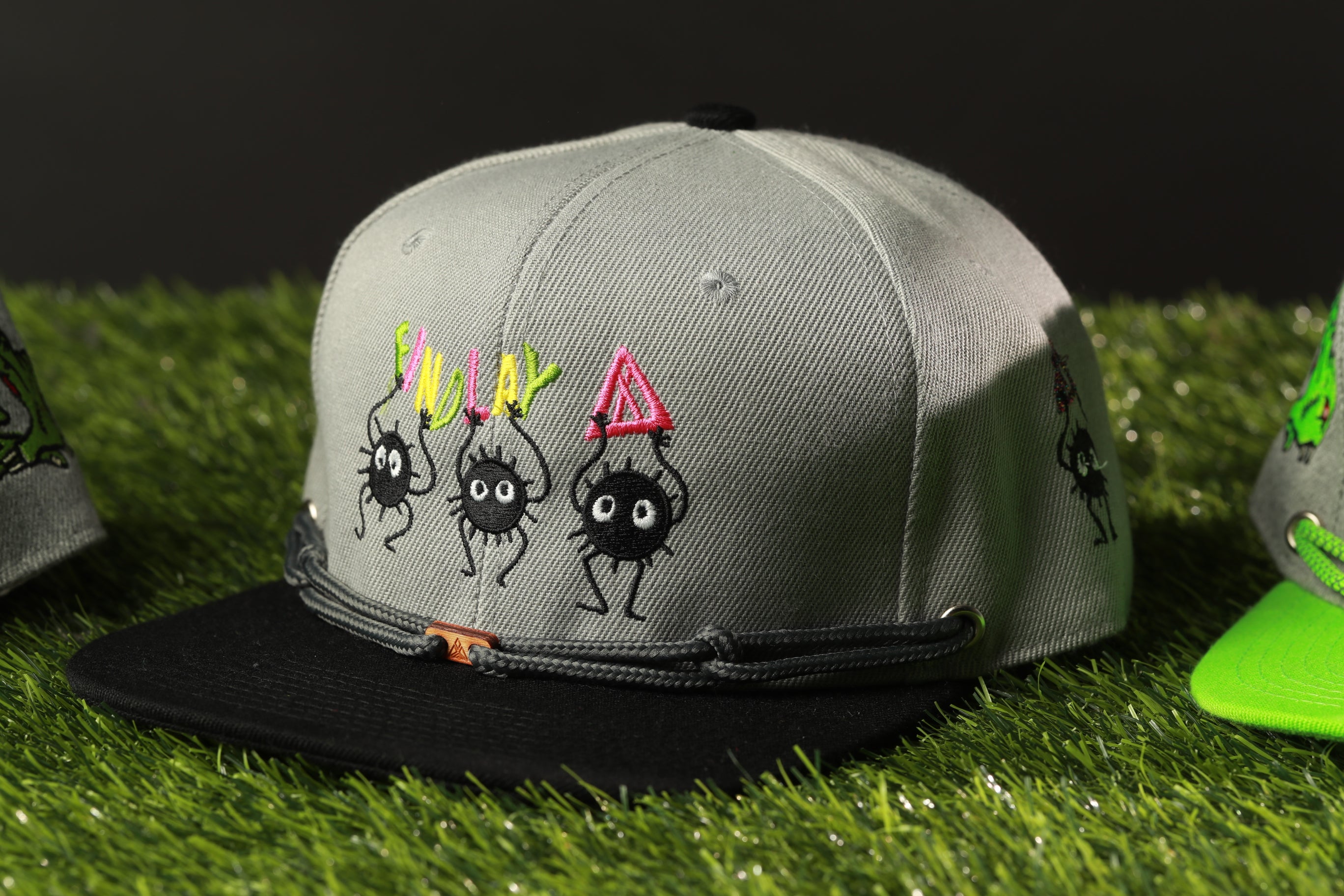 A gray baseball cap with a black brim and a black band, featuring three cartoon spider characters with colorful accents.