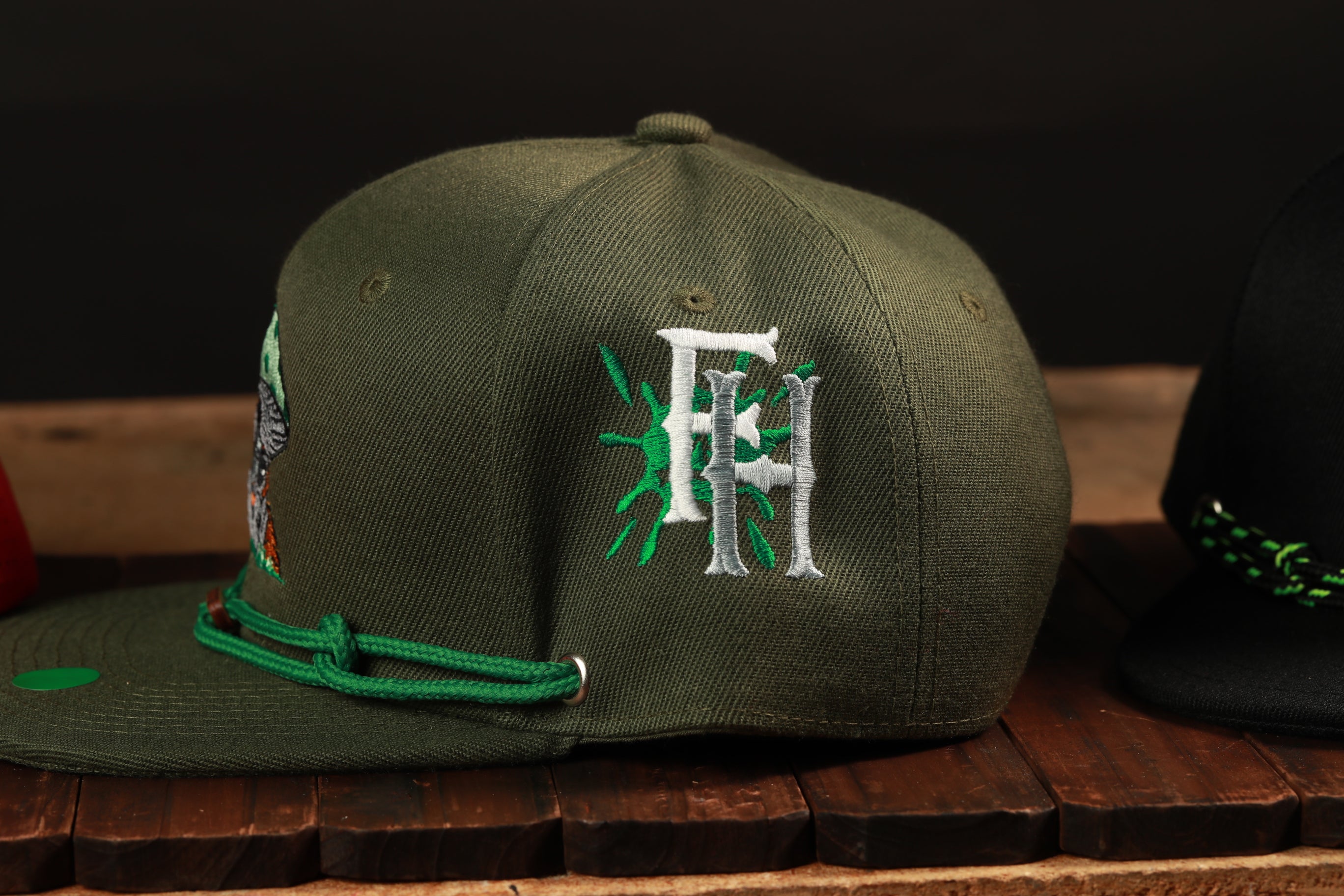 A close-up view of a green baseball cap with a white embroidered logo on the side, displayed on a wooden surface.