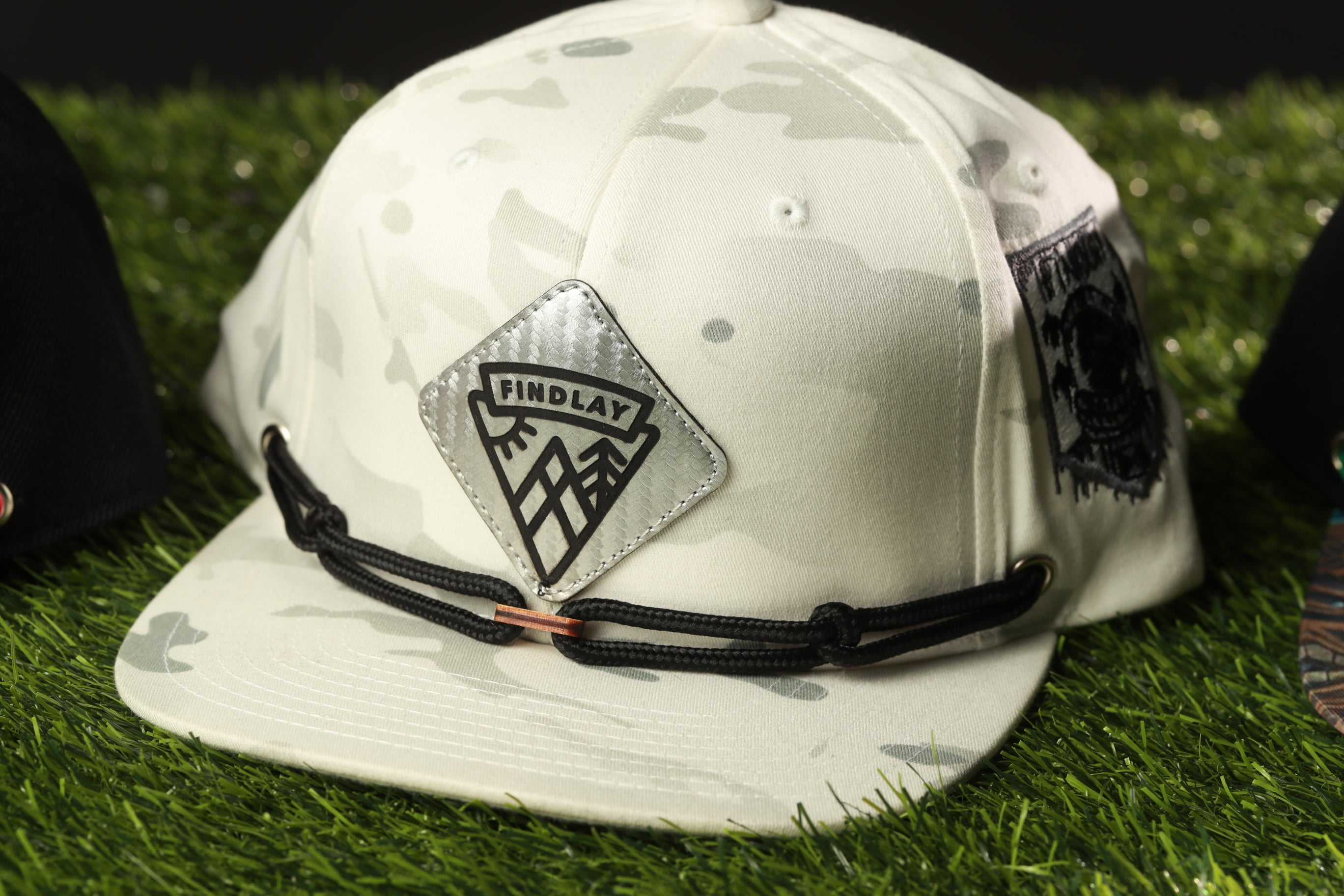 A white camouflage baseball cap with a black and silver logo on the front, placed on a grassy surface.