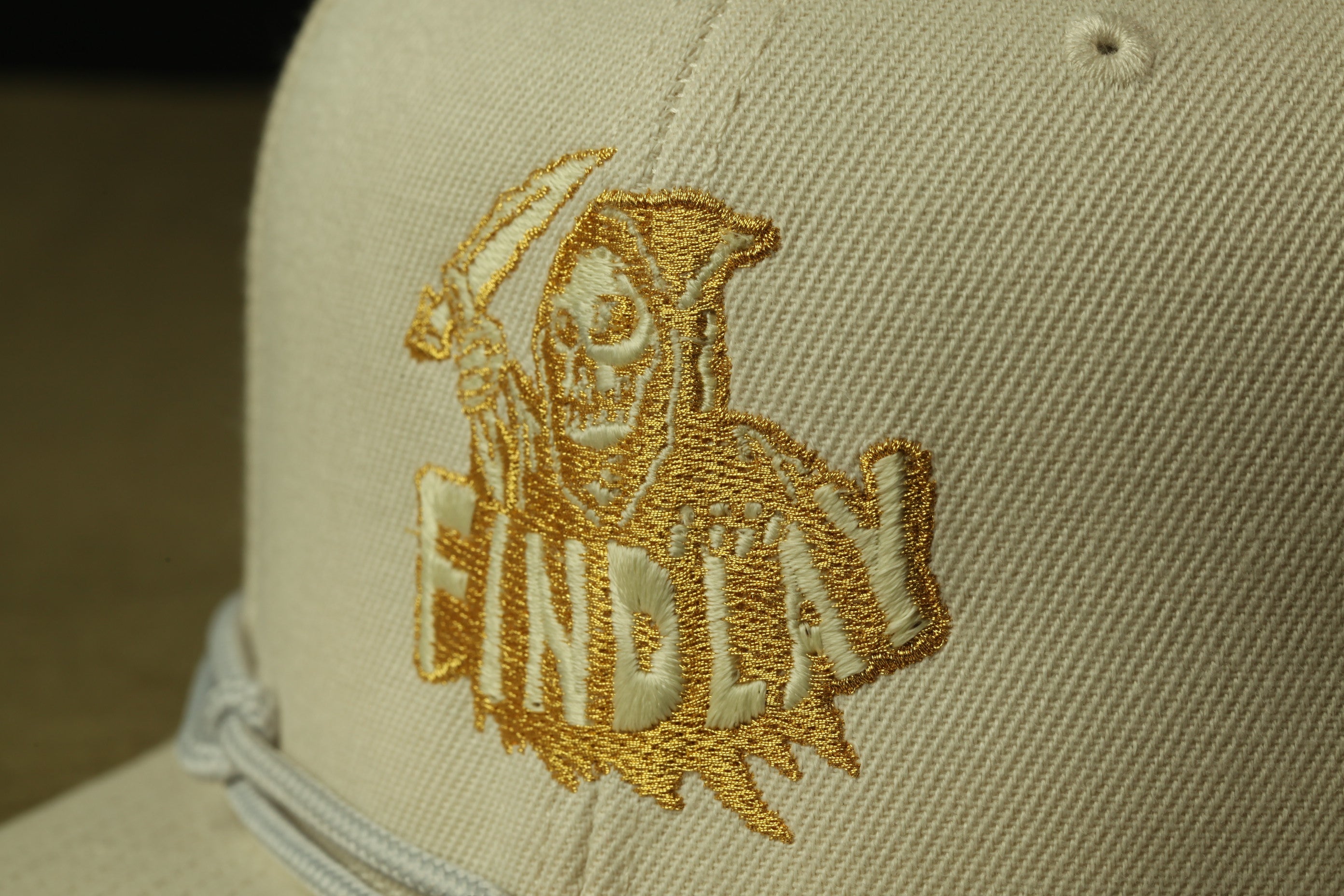 A close-up view of a beige baseball cap with a gold embroidered logo on the front.