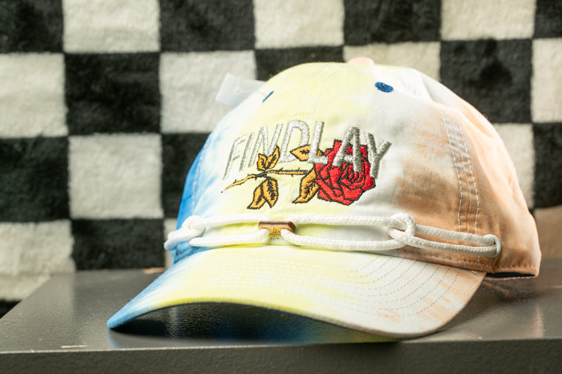A tie-dye baseball cap with the word "FINDLAY" embroidered on it, sitting on a surface against a checkered background.