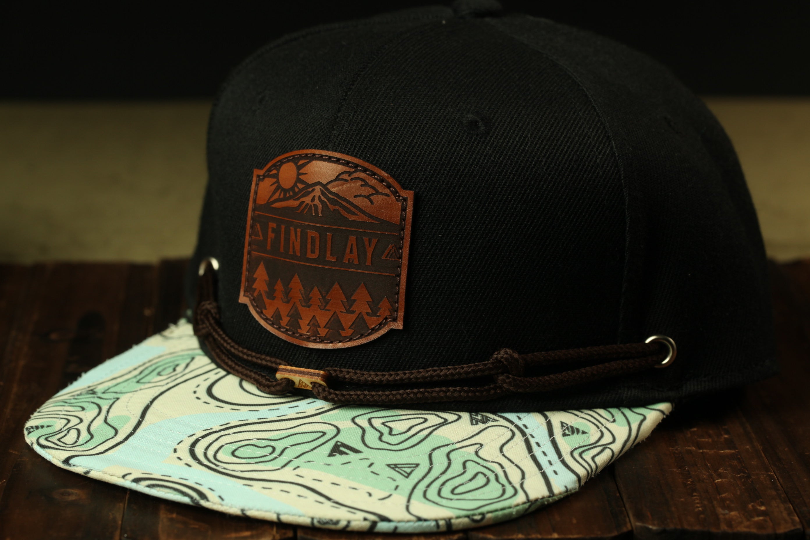 A black baseball cap with a leather patch on the front, featuring a mountain landscape design and the word "FINDLAY" printed on it.