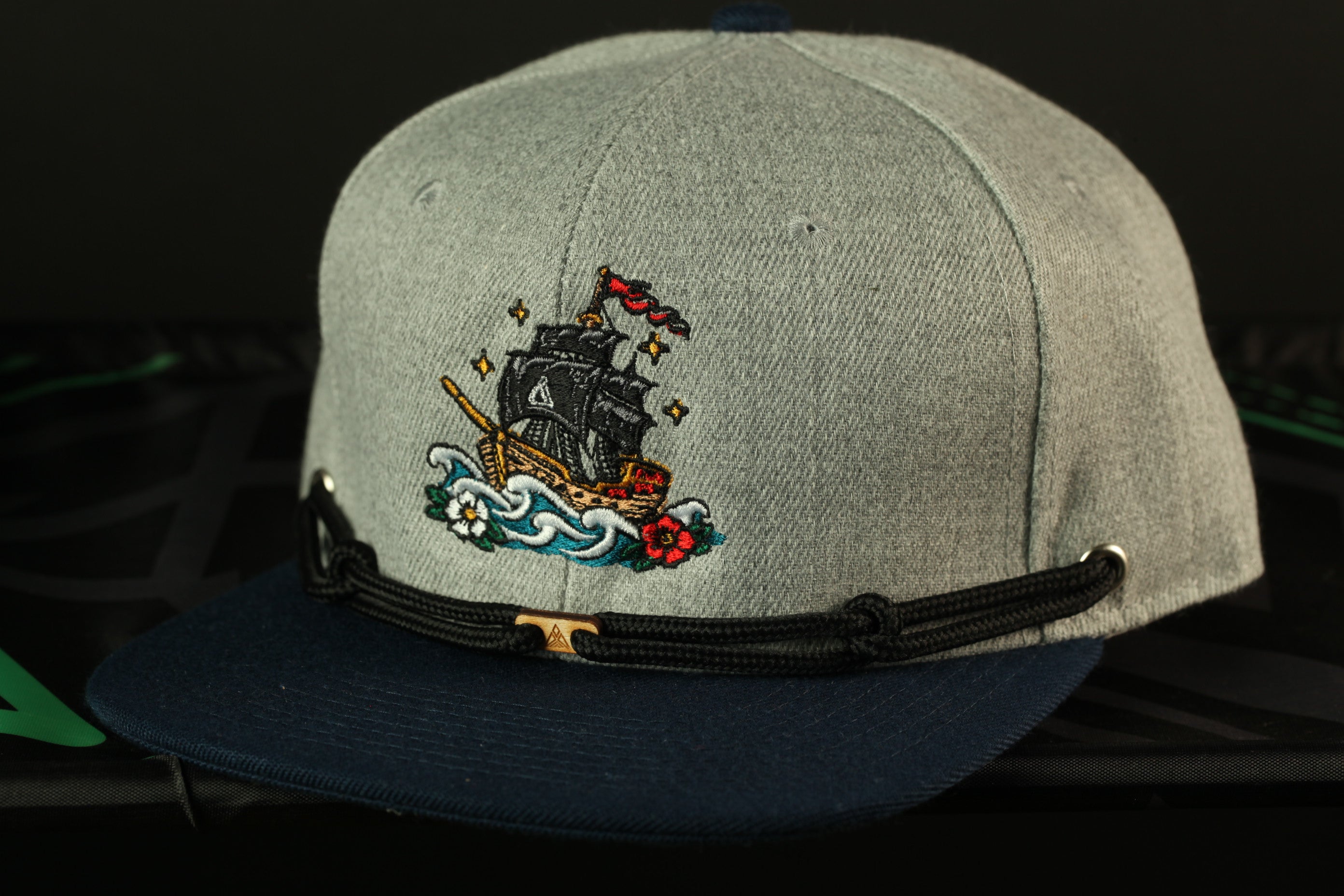 A gray baseball cap with a black brim and a navy blue band, featuring an embroidered image of a ship sailing on a wave.