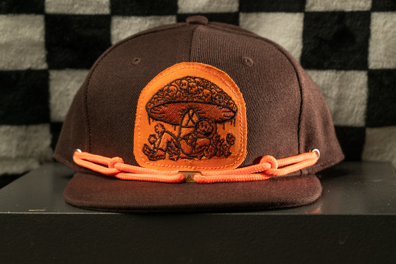 A brown baseball cap with an orange patch featuring a mushroom design and orange straps.
