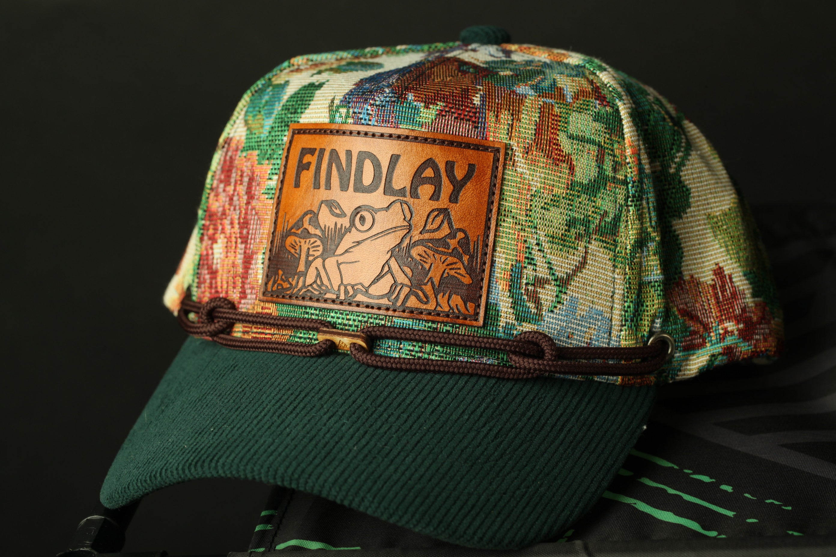 A colorful, patterned baseball cap with a frog design and the word "FINDLAY" printed on it.
