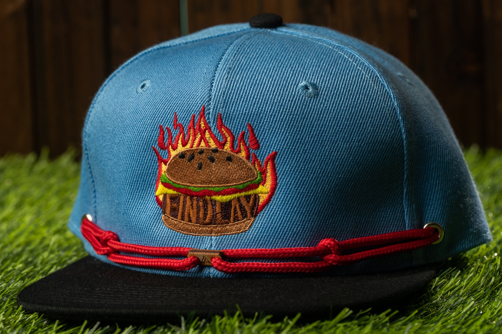 A blue baseball cap with a red string and a graphic of a hamburger on it, placed on a grassy surface.
