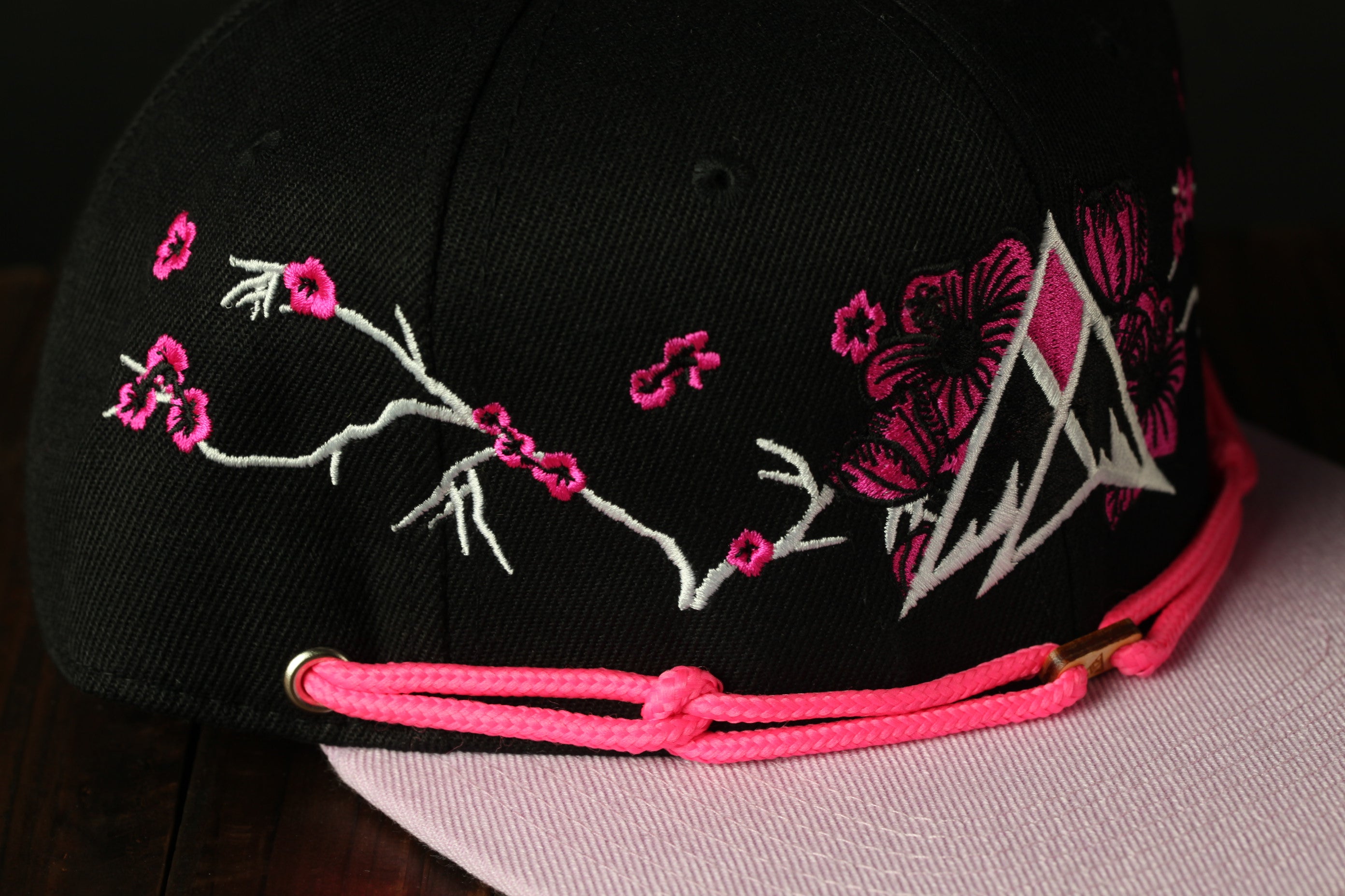 A close-up view of a black baseball cap with pink floral embroidery and a pink braided strap.
