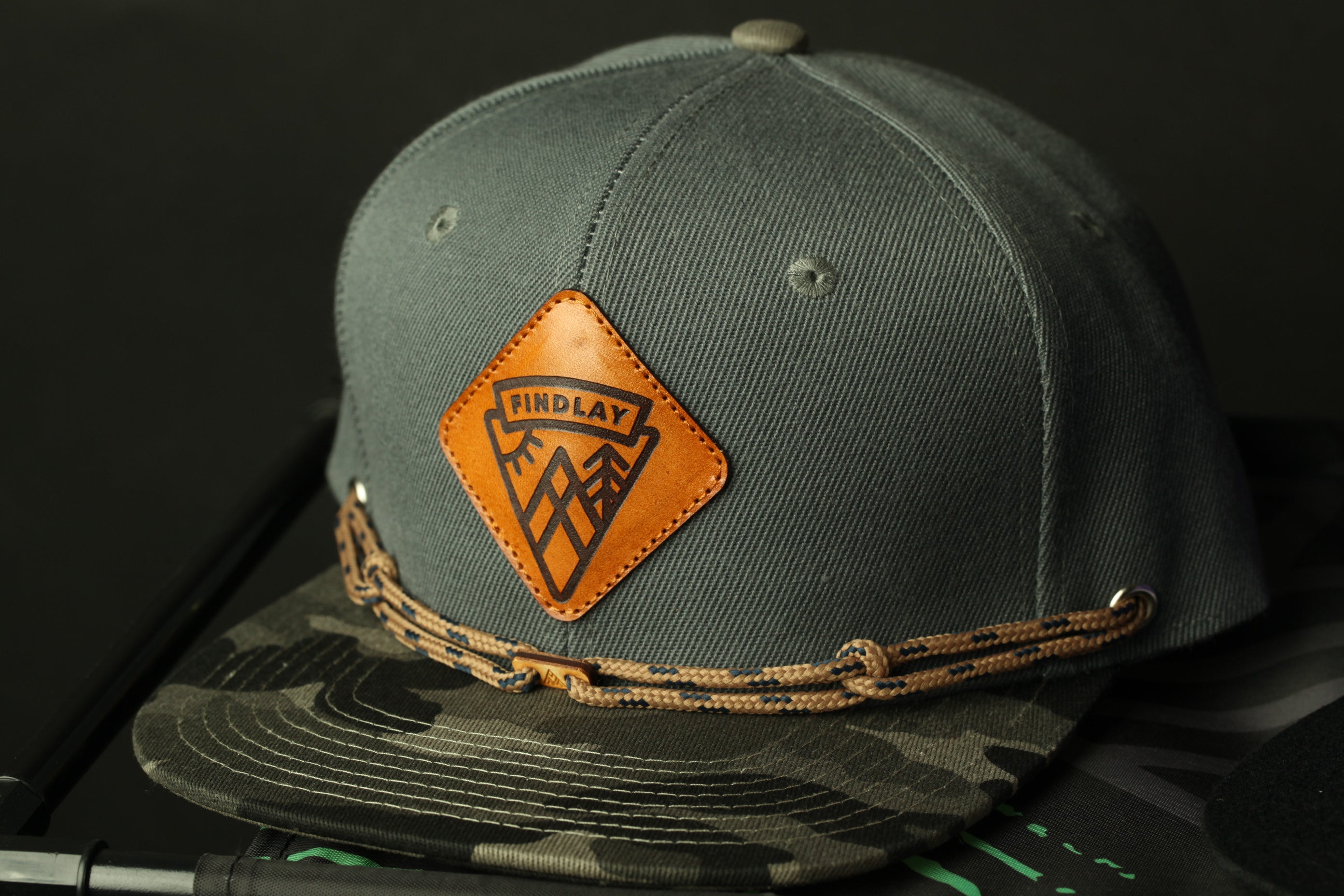 A gray baseball cap with a camouflage pattern and a leather patch on the front, featuring the text "FINDLAY" and a mountain silhouette.