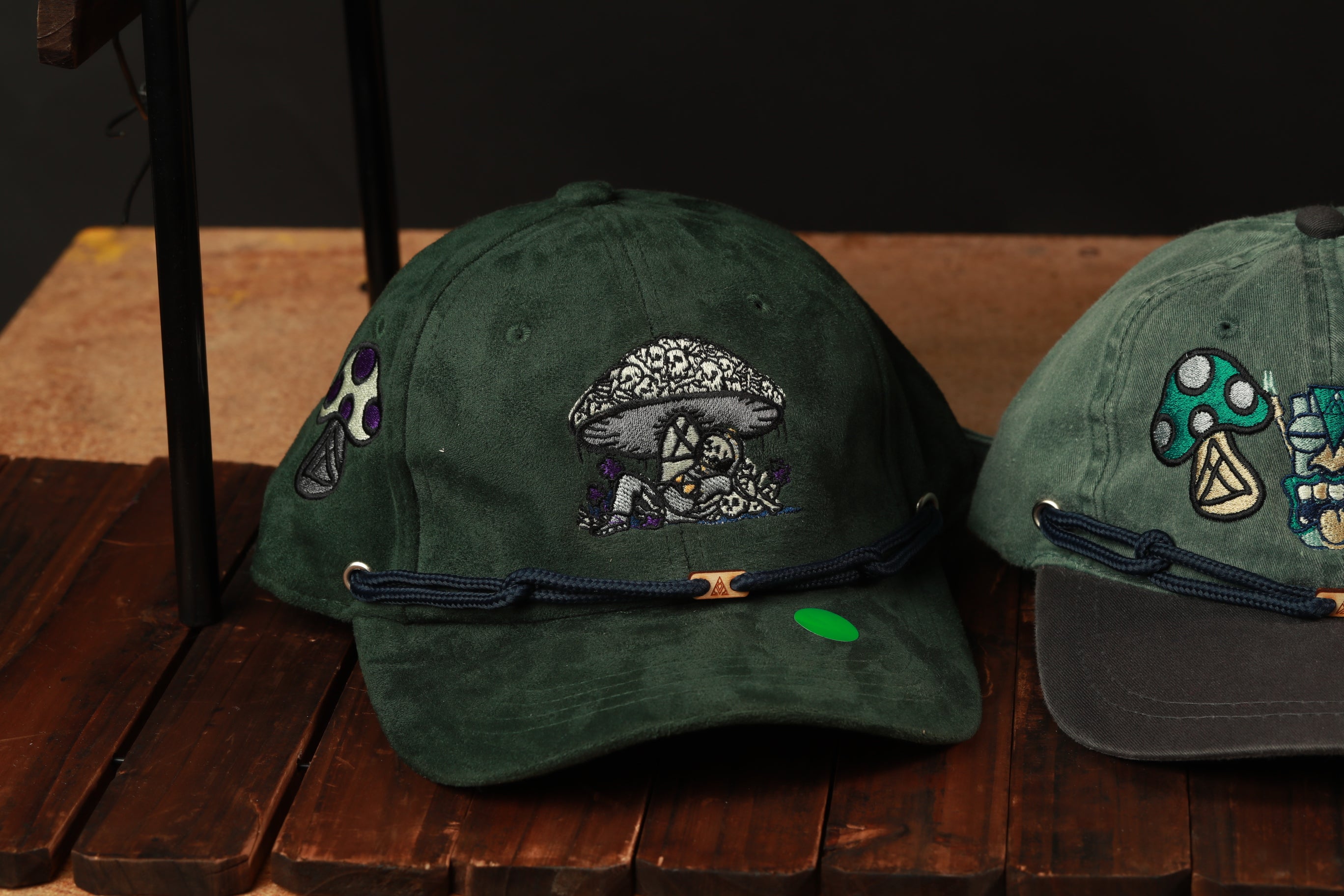Two green baseball caps with mushroom designs, one with a green tag attached to it, are displayed on a wooden surface.