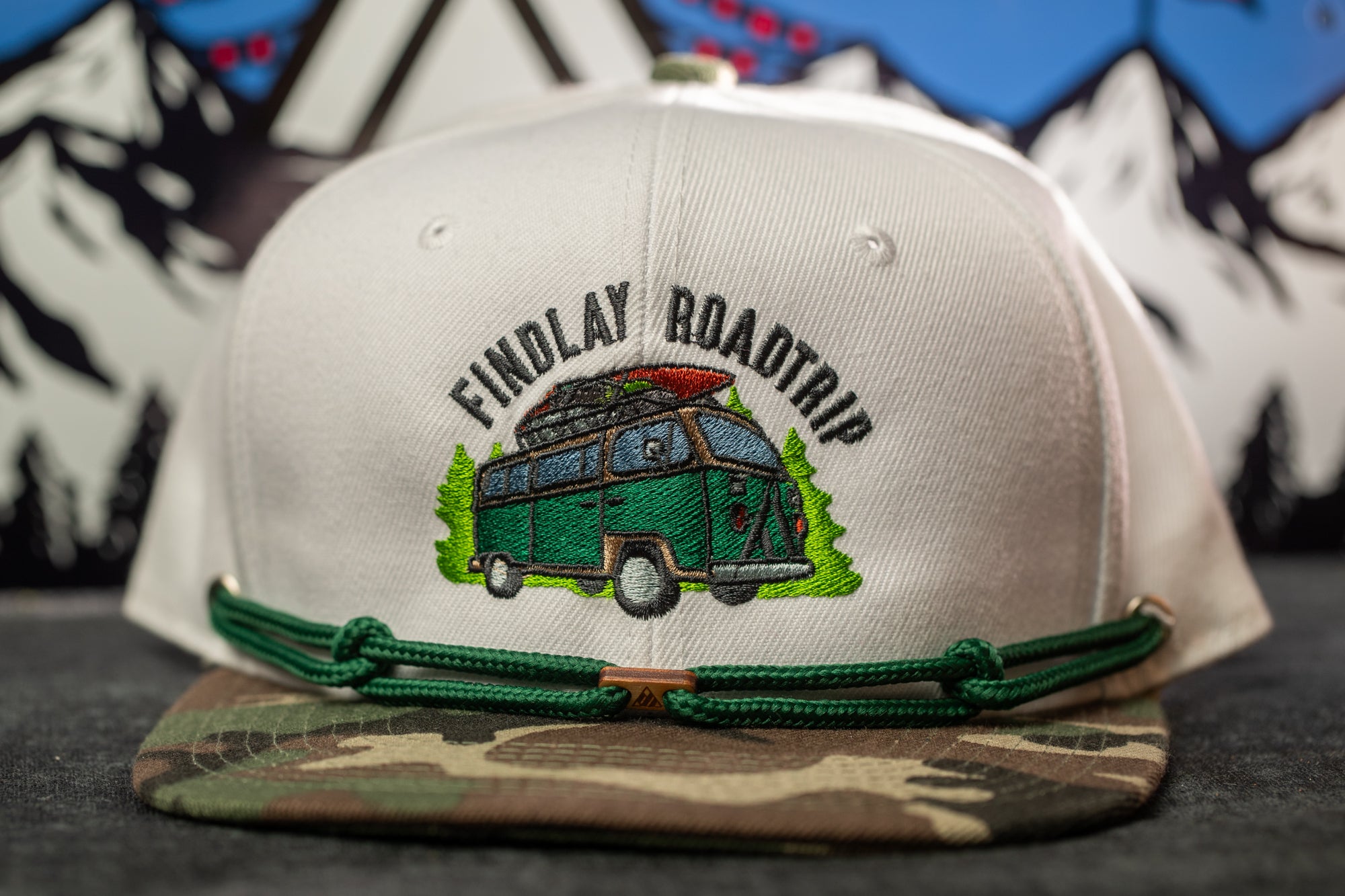A white baseball cap with a green camo pattern and a graphic of a camper van, along with the text "FINDLAY ROADTRIP".