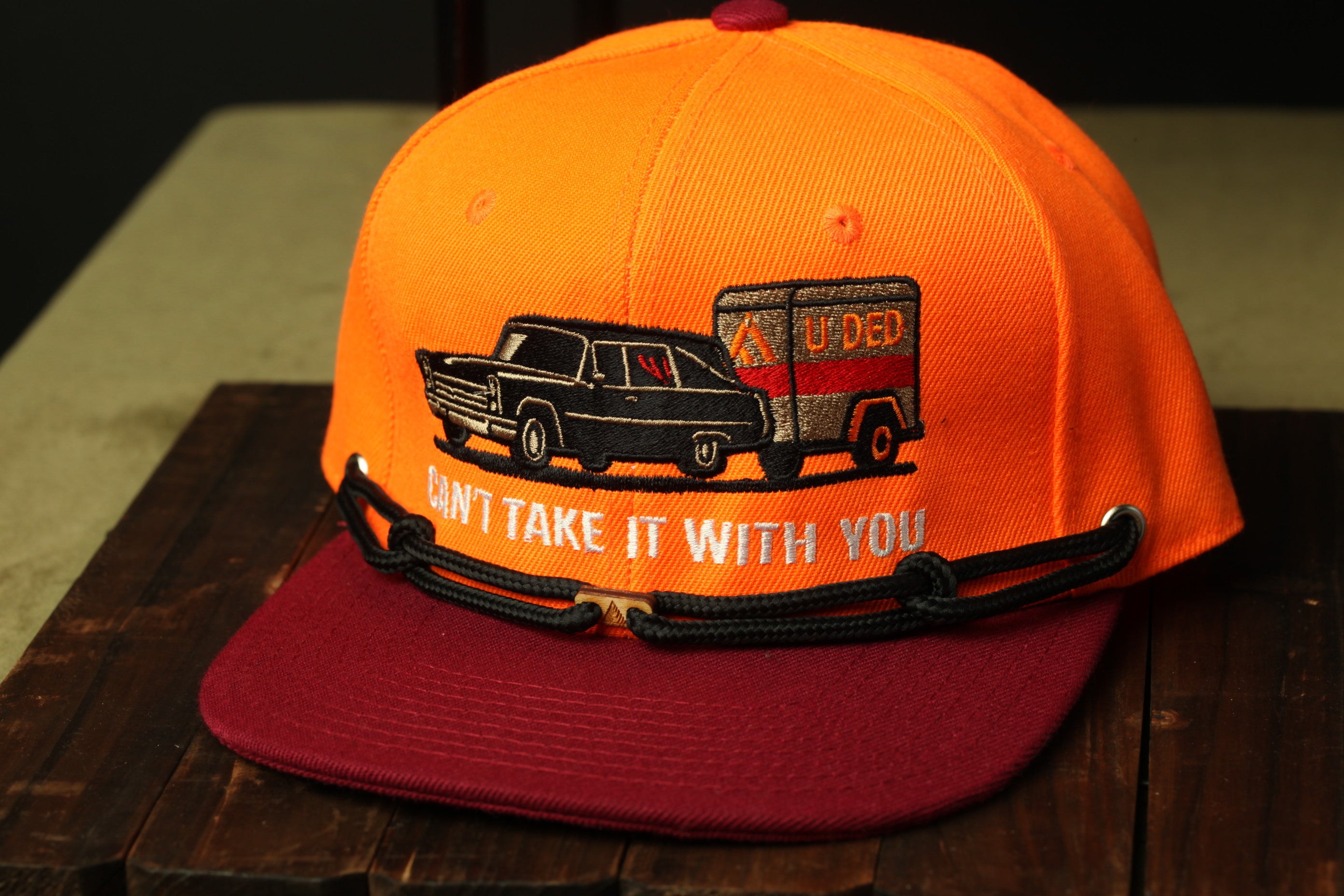 An orange baseball cap with a graphic design depicting a car and a trailer, along with the text "CANT TAKEOW IT WITH YOU".