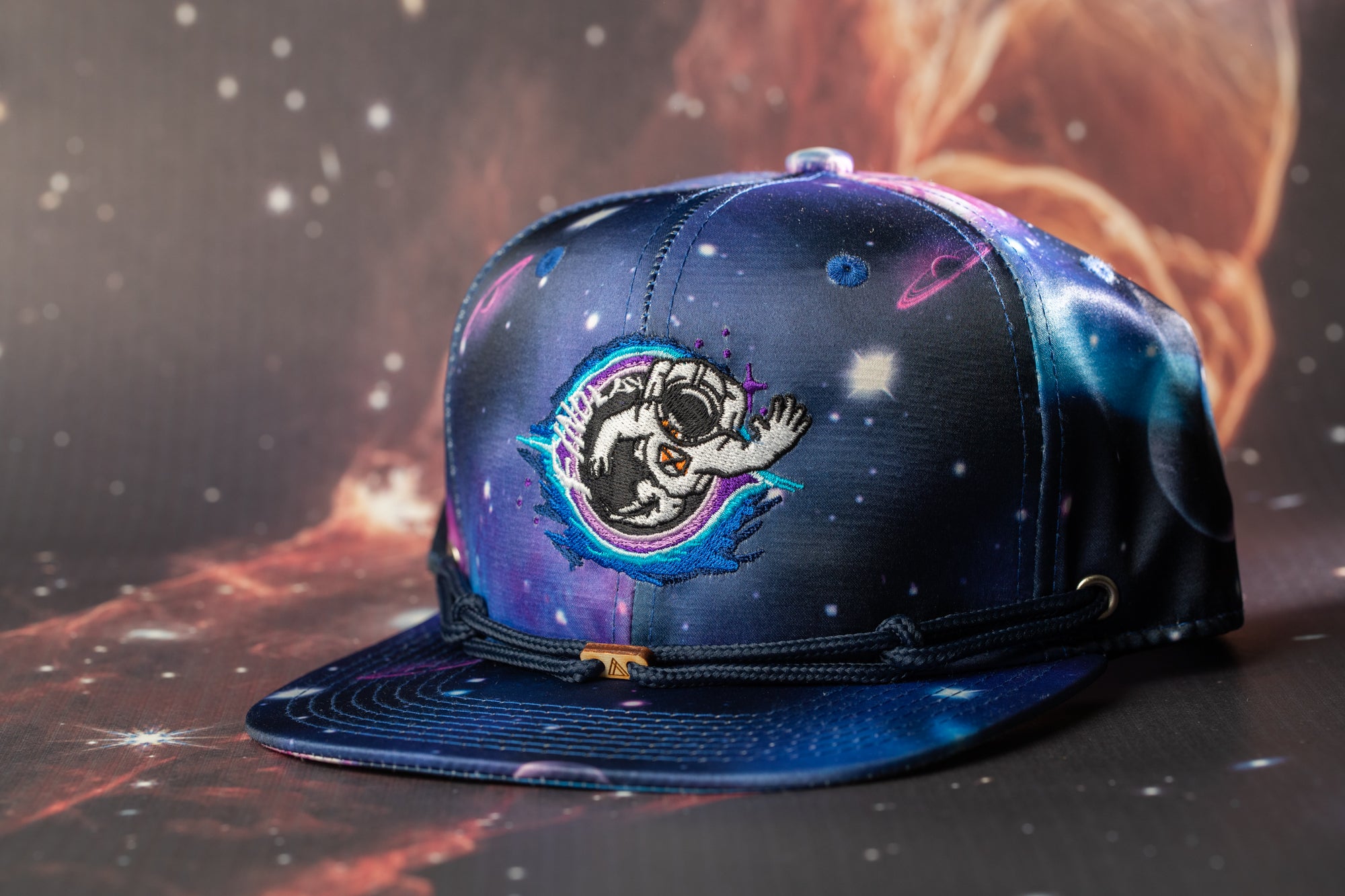 A colorful baseball cap with a space-themed design, featuring a cartoon astronaut or alien character, is displayed against a backdrop of a starry night sky.