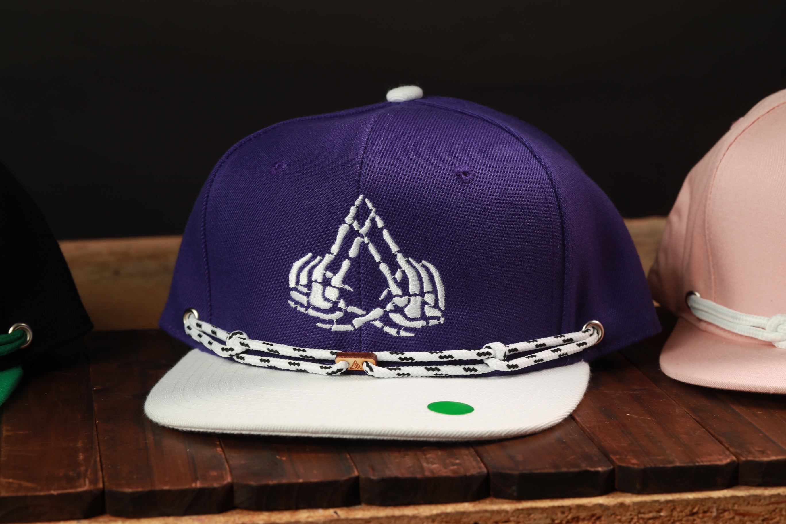 A purple baseball cap with a white skull and crossbones design, featuring a white lace-like strap, is displayed on a wooden surface.