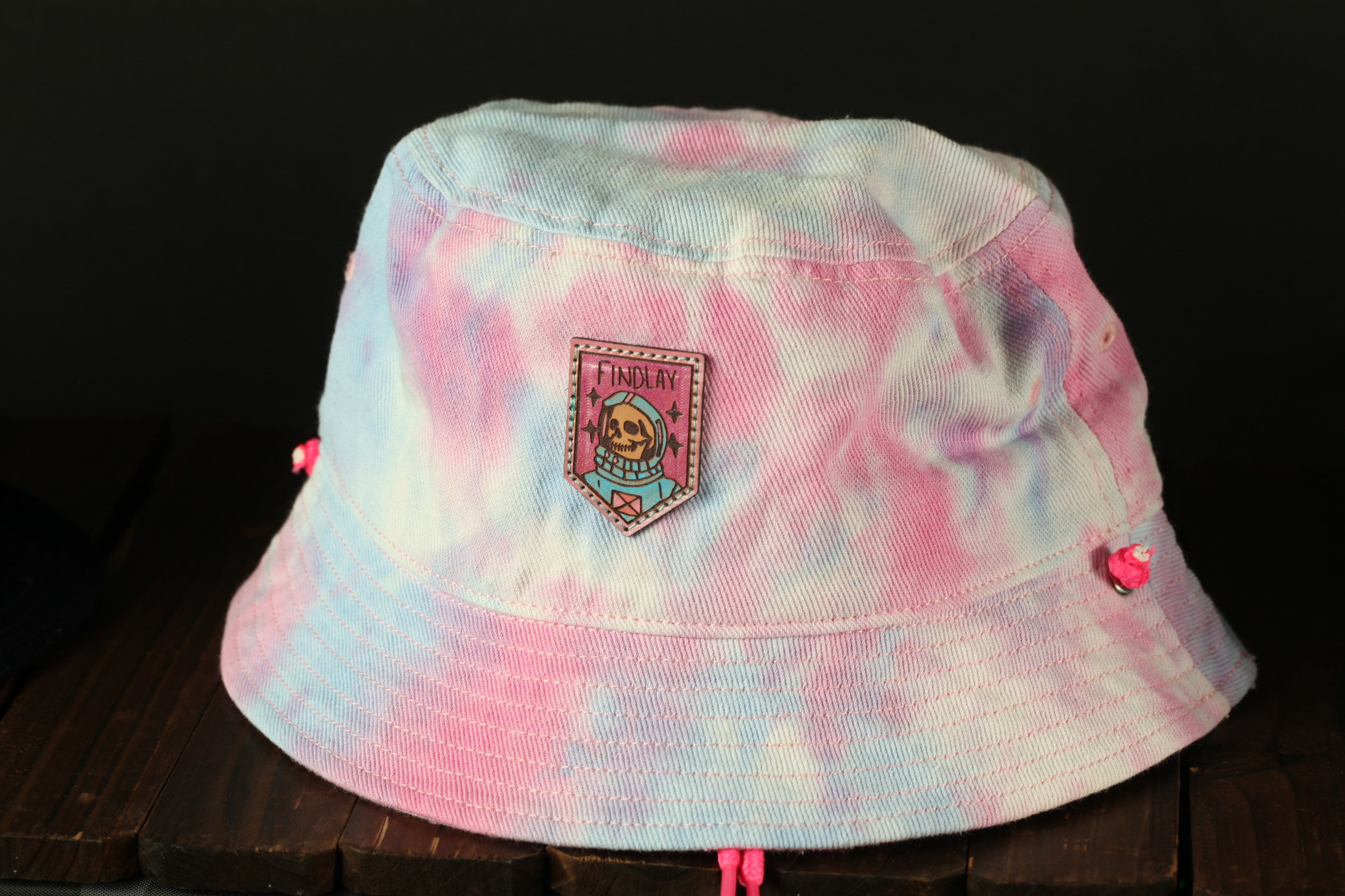 A pink and blue tie-dye bucket hat with a skull and crossbones patch on the front, sitting on a wooden surface.