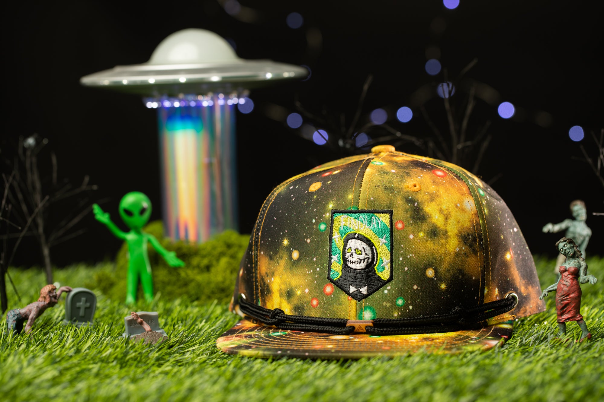 a colorful baseball cap with a space-themed design, surrounded by various small figurines and a spaceship in the background.