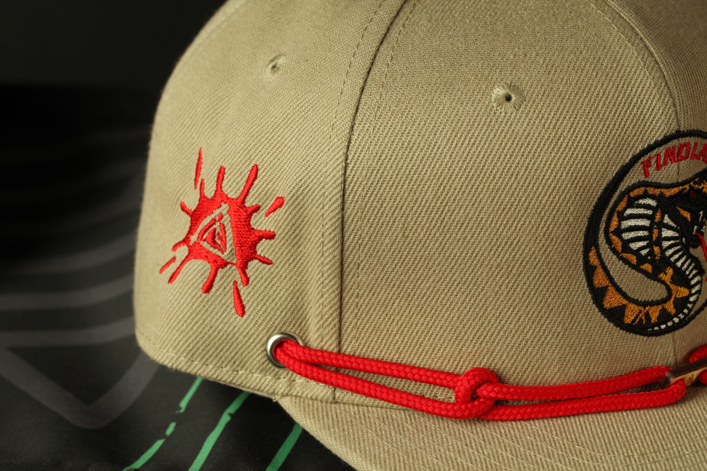 A beige baseball cap with a red splatter design and a snake logo on the front.