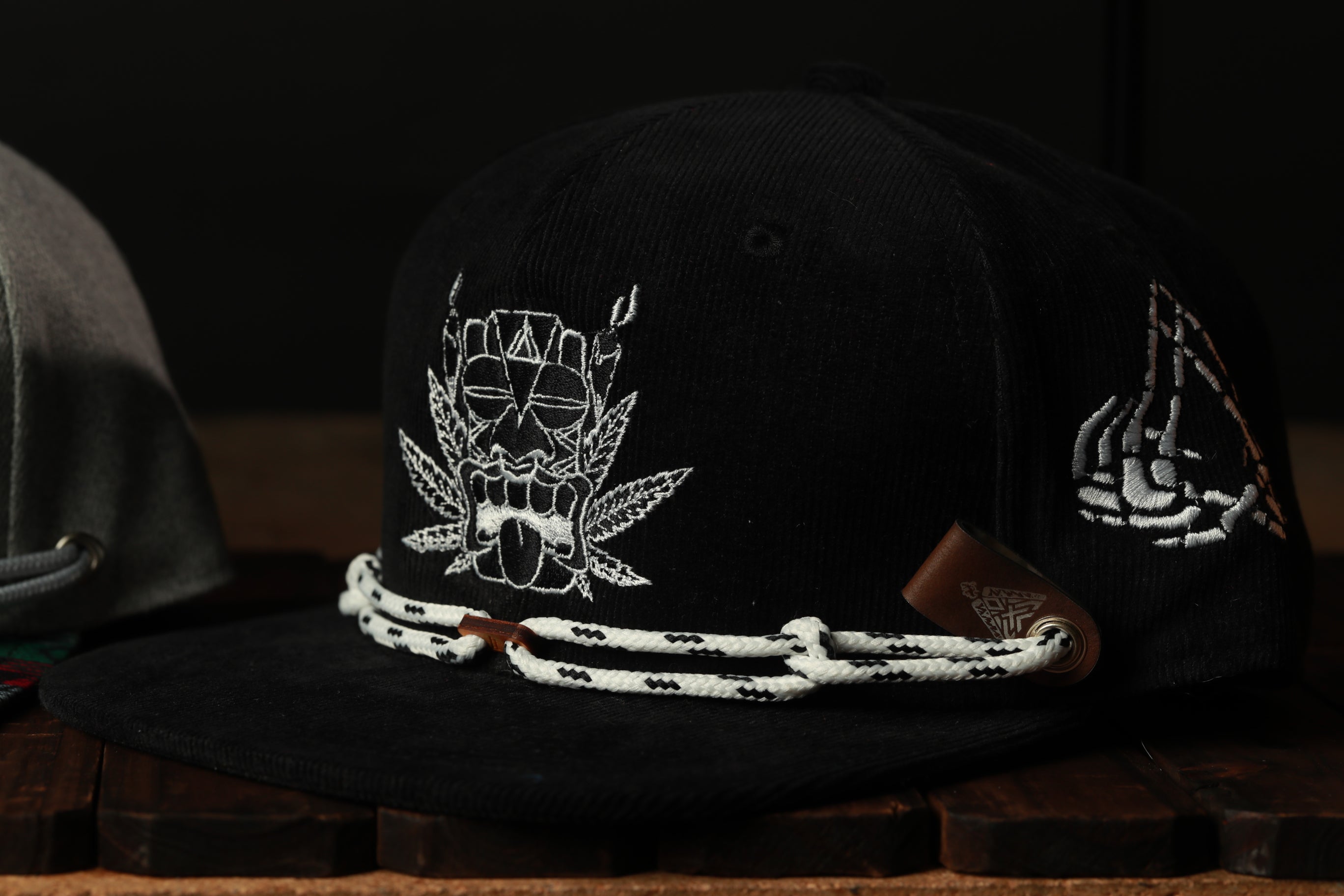 A black baseball cap with white embroidered designs, including a skull and crossbones, and a white lace-like strap.