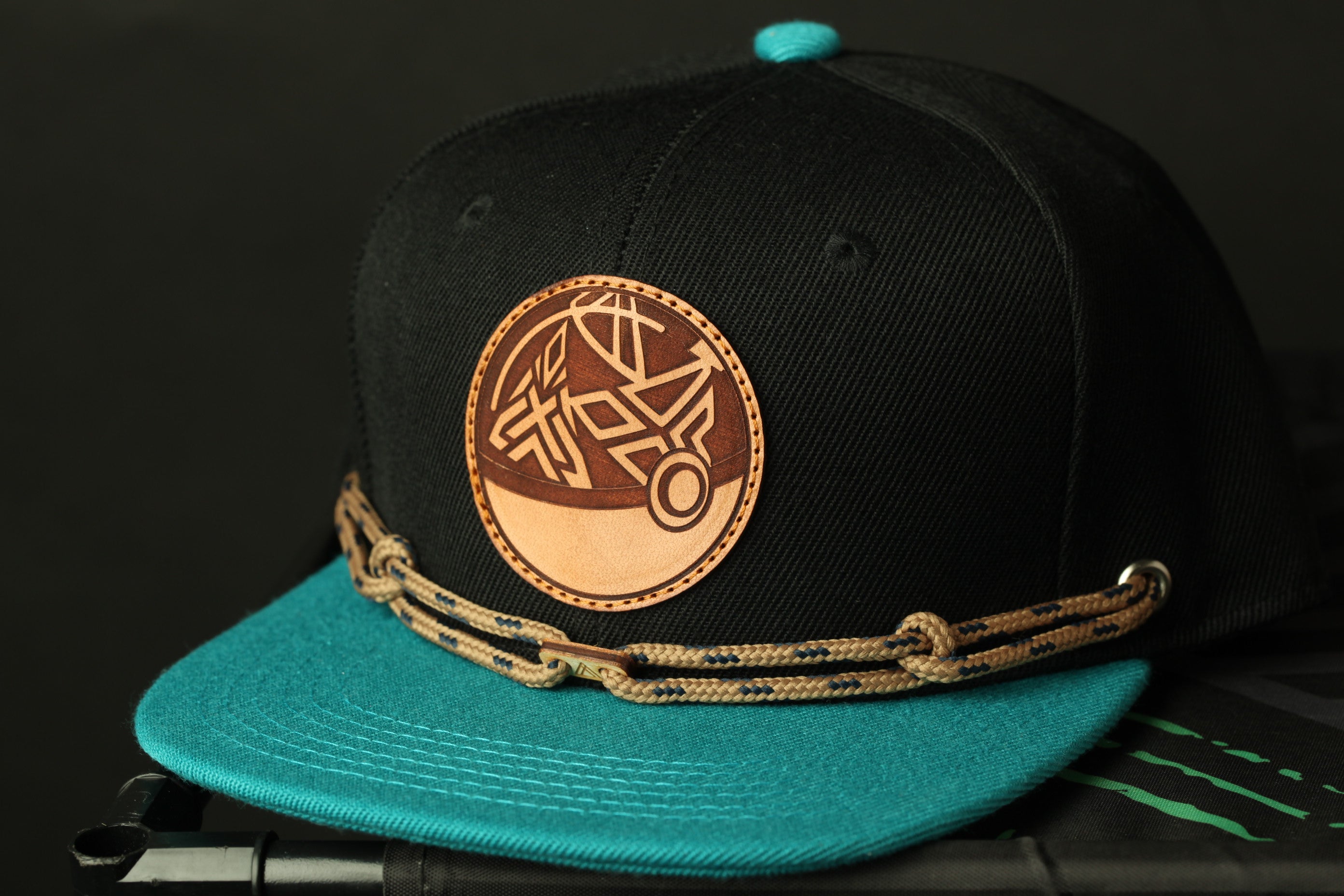 A black baseball cap with a teal brim and a brown leather patch on the front, featuring a circular design.