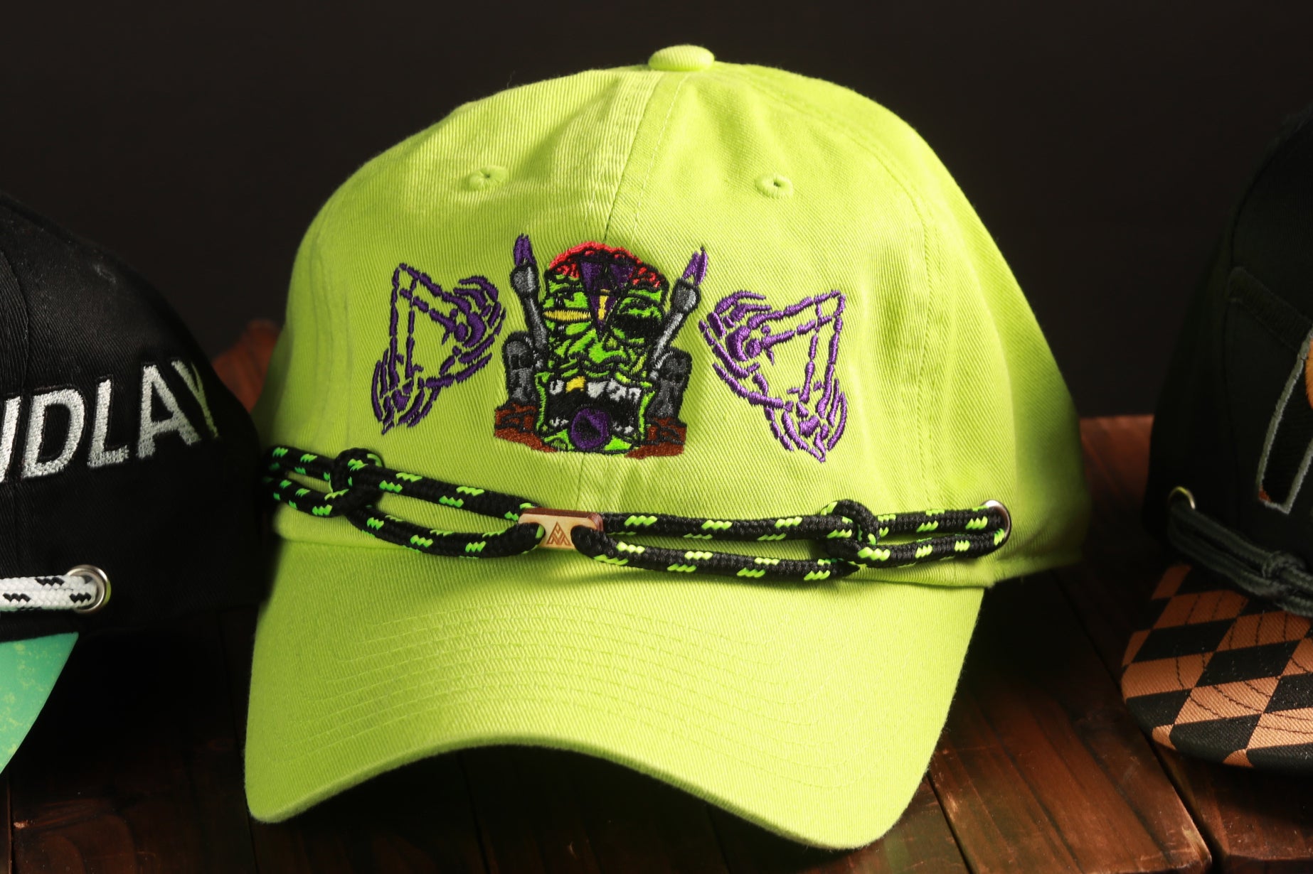 A bright yellow baseball cap with a unique design featuring a green monster-like creature and purple geometric shapes.