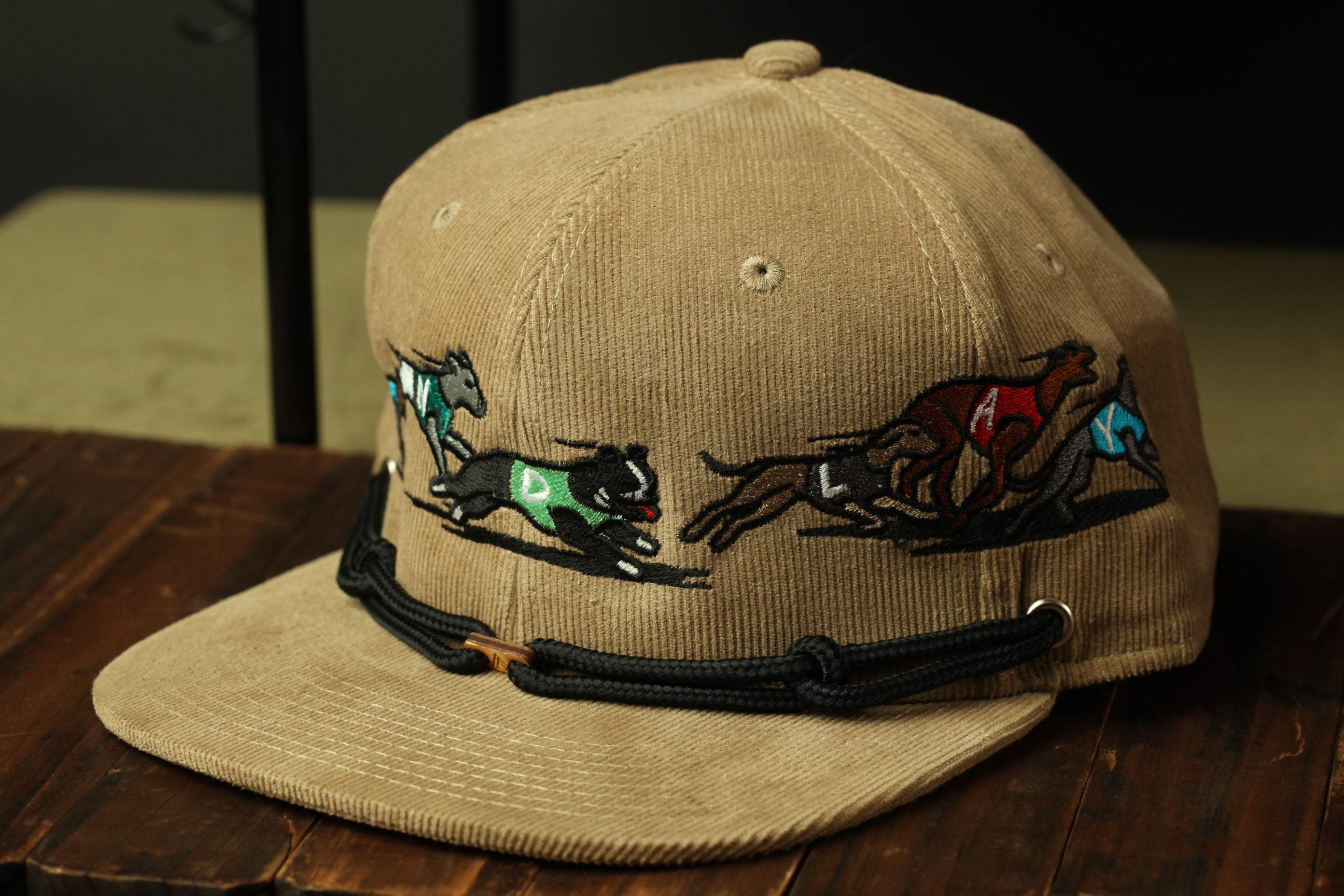 A beige baseball cap with a black strap and embroidered designs of dogs running.