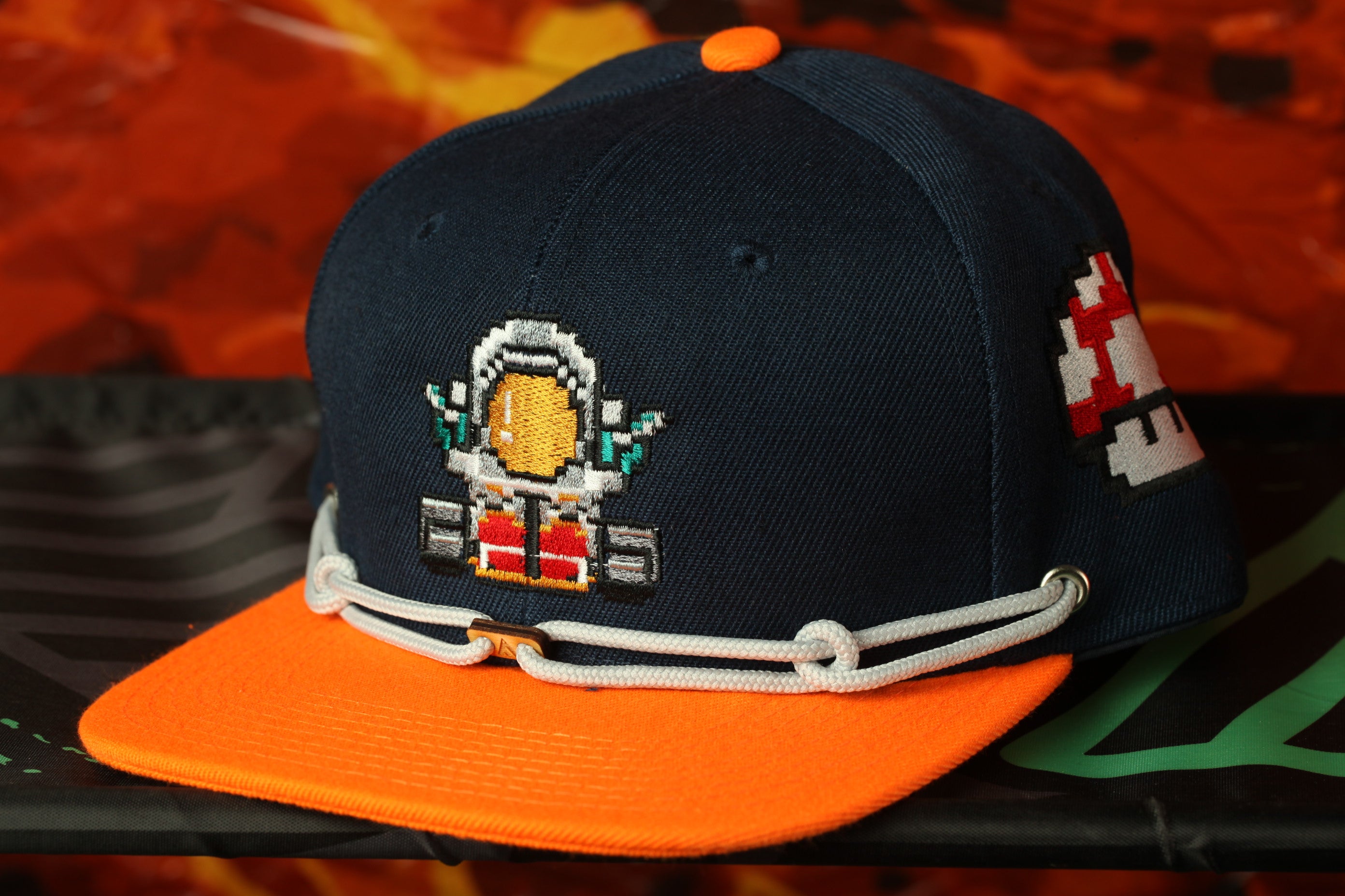 A navy blue baseball cap with an orange brim and a pixelated character design on the front.